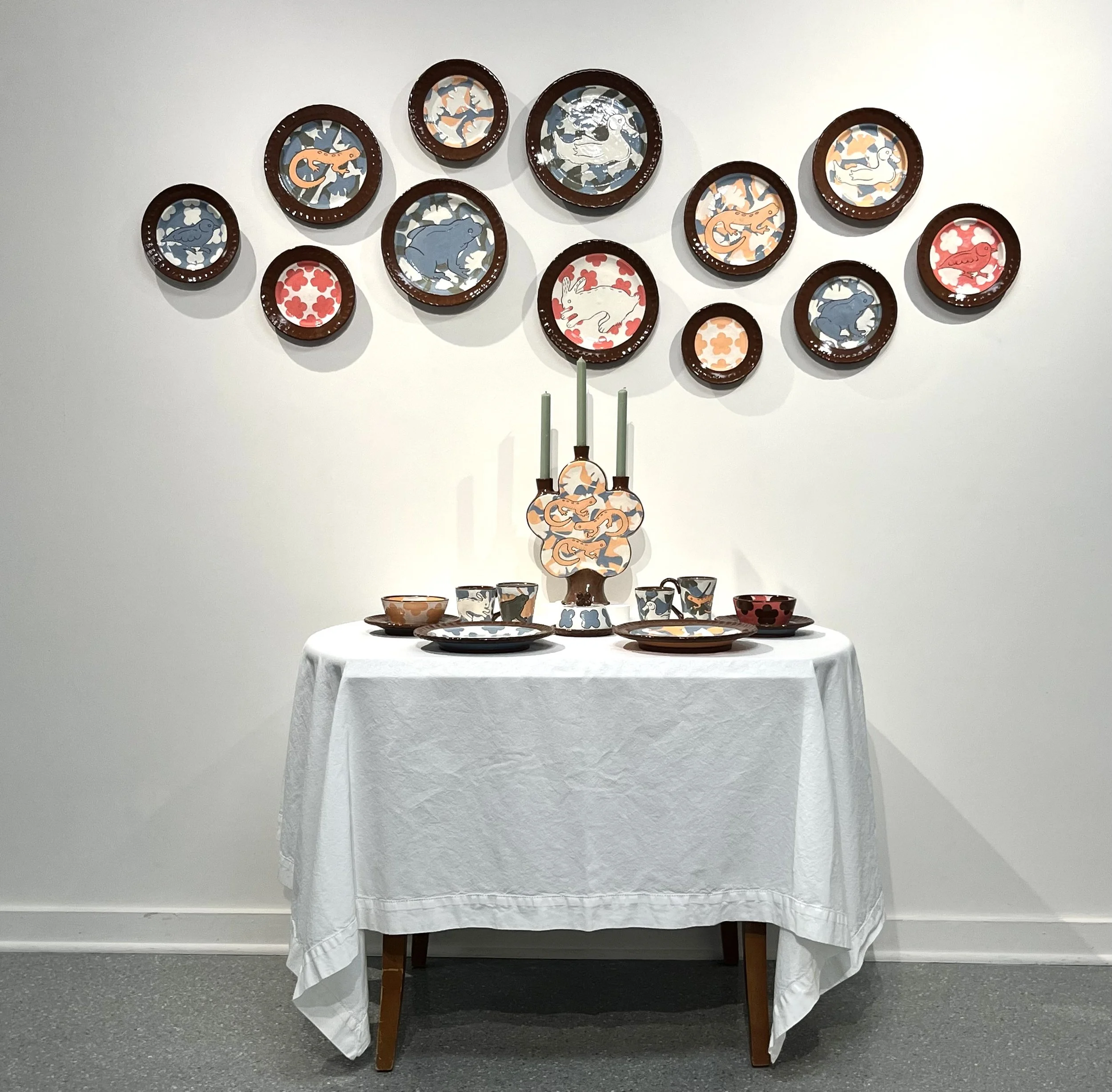A table with a white tablecloth displaying various ceramic dishes and bowls with animal motifs, arranged beneath a wall hanging of decorated plates featuring animal designs.