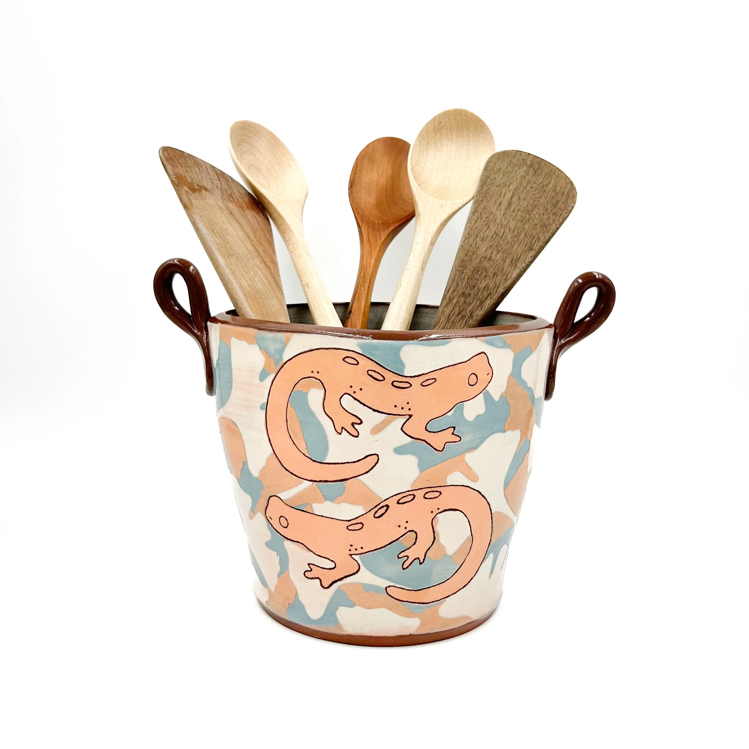 A ceramic cup decorated with a cartoon dinosaur pattern, holding wooden utensils.