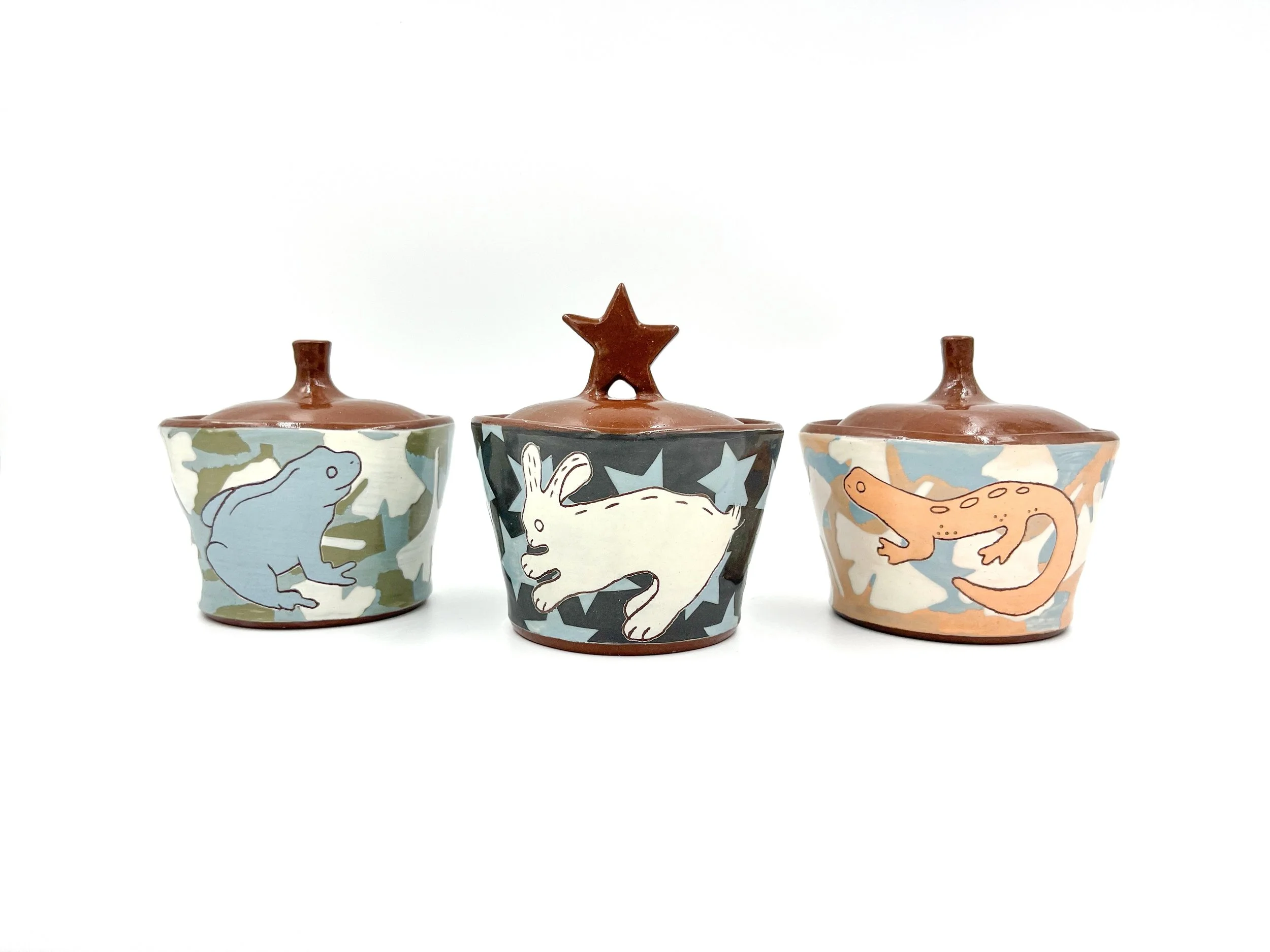 Set of three ceramic cups with colorful dinosaurs and star patterns on white backgrounds and brown lids topped with star and dinosaur figures.