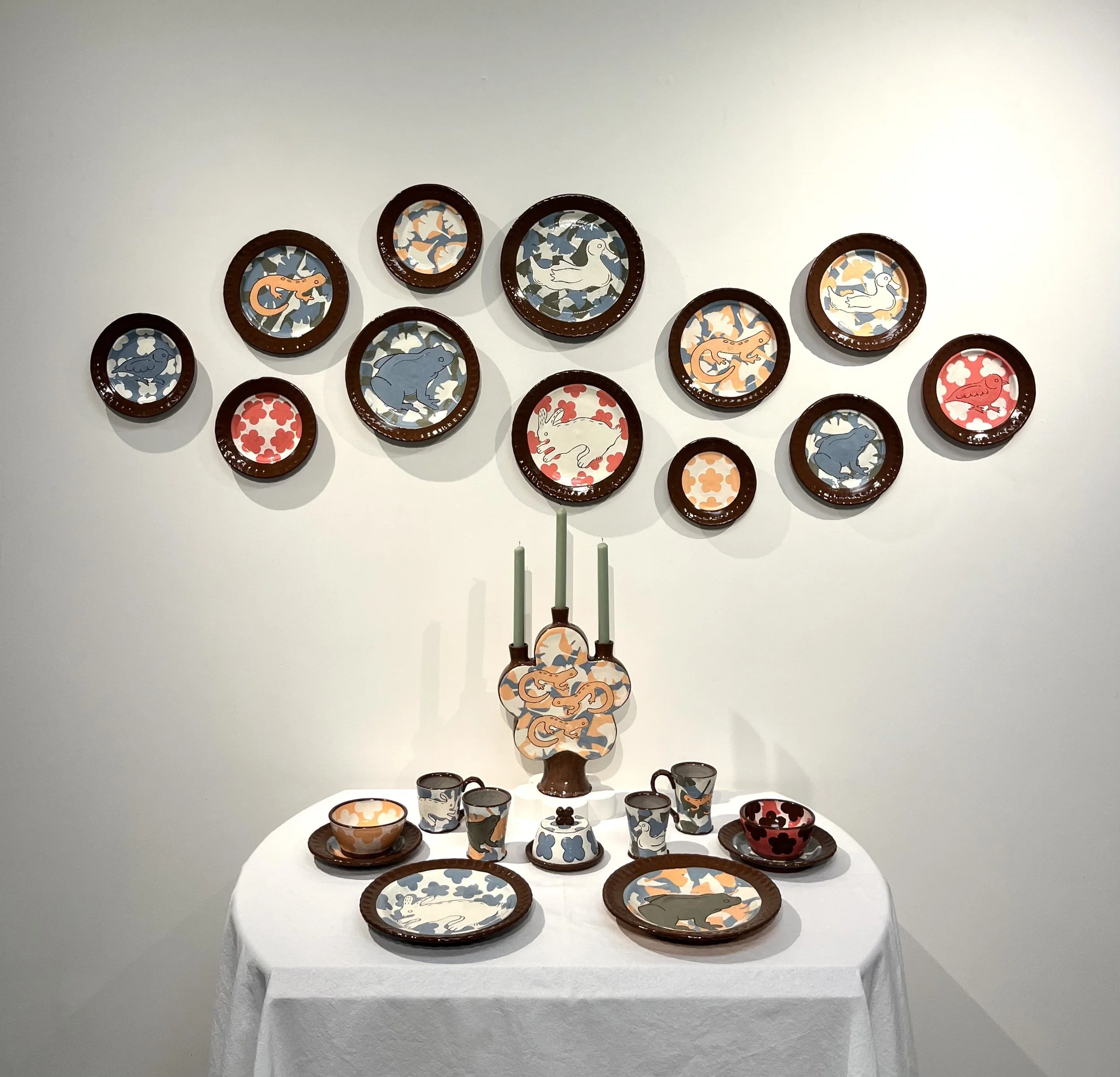 Display of ceramic dishes and cups with animal and floral patterns, arranged on a three-tiered stand and shelves against a white wall.