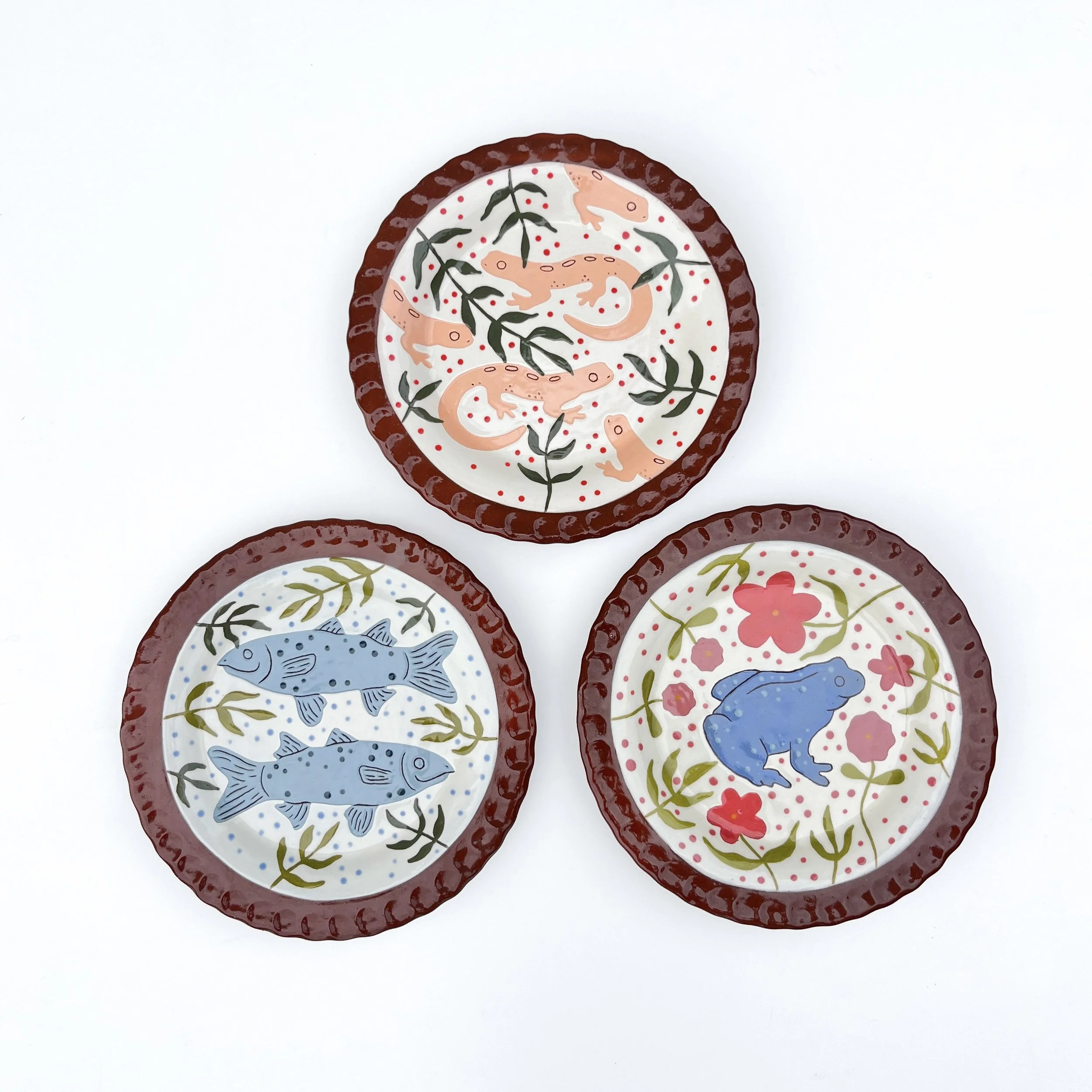 Decorative ceramic plate with a design of pink salamanders, green leaves, and red dots on a white background, with a red scalloped border.