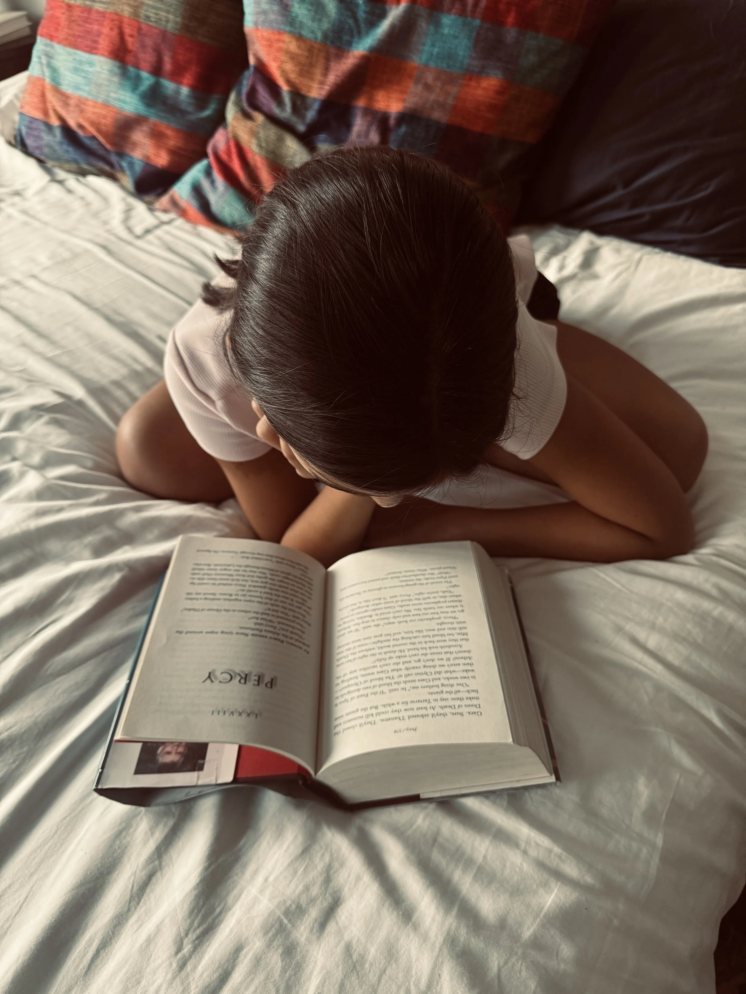 Person with dark hair reading a book titled 'PEAKY' on a white bed with a colorful plaid pillow.