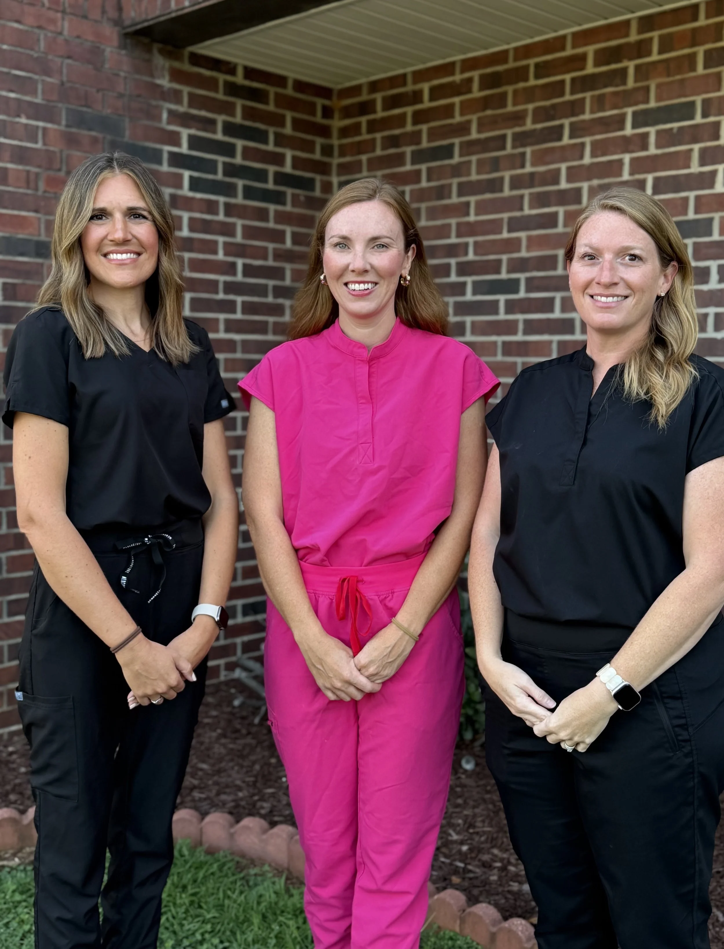 Three women standing outdoors in front of a brick wall, smiling at the camera. The woman in the middle is wearing a pink scrub top, while the women on either side are wearing black scrubs.