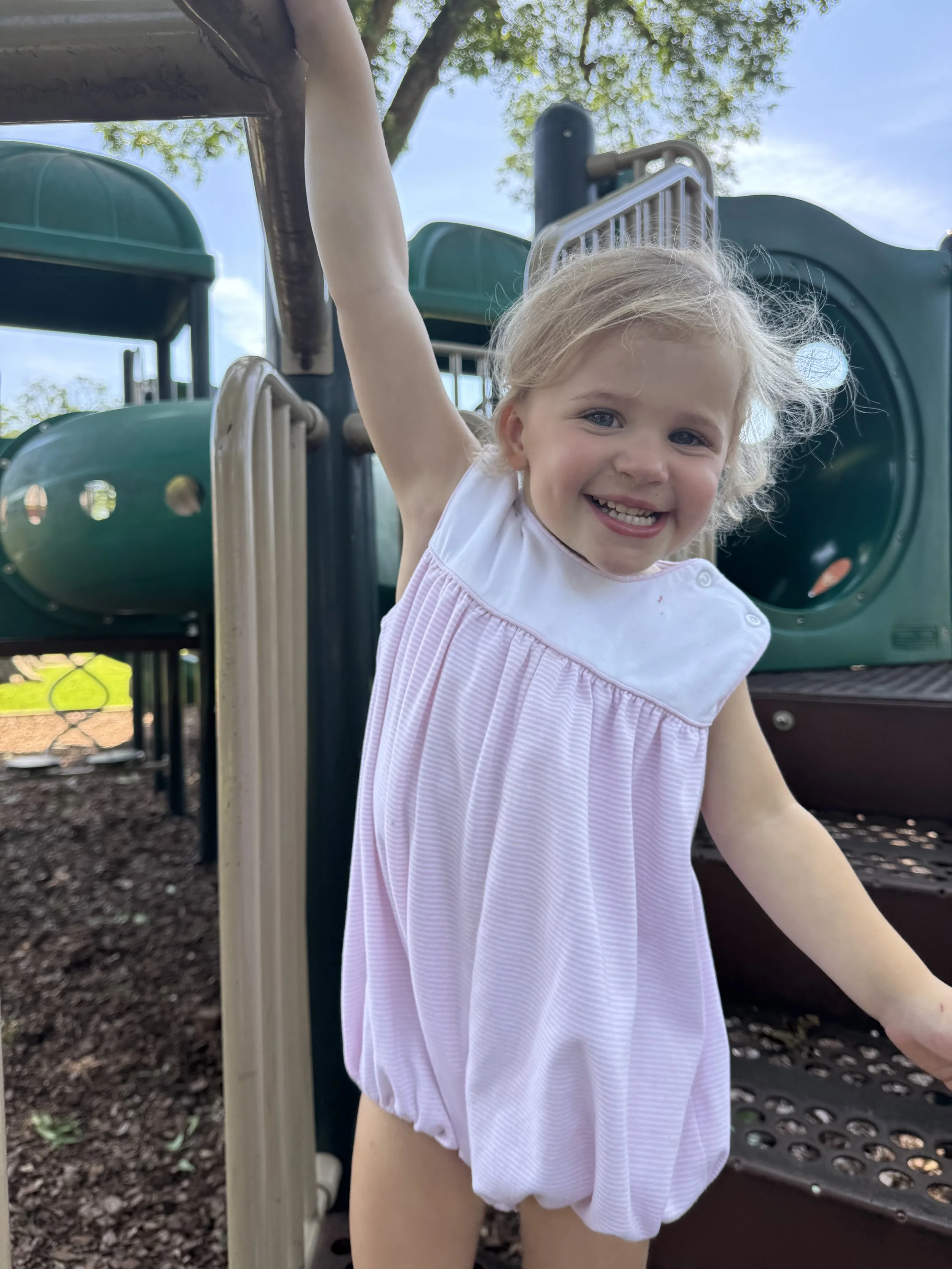 A young girl in a light pink and white dress playing on playground equipment outdoors in a park, smiling at the camera with a blue sky and trees in the background.
