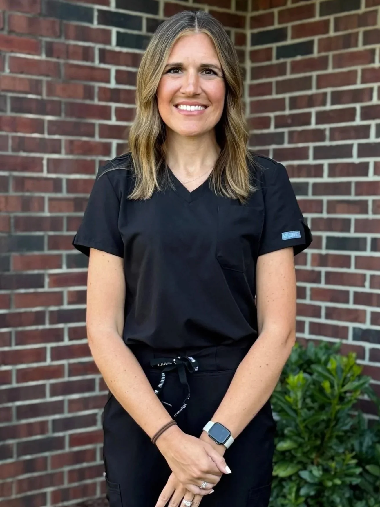 A woman with shoulder-length light brown hair, smiling, wearing a black medical uniform and standing outside in front of a brick wall and greenery.