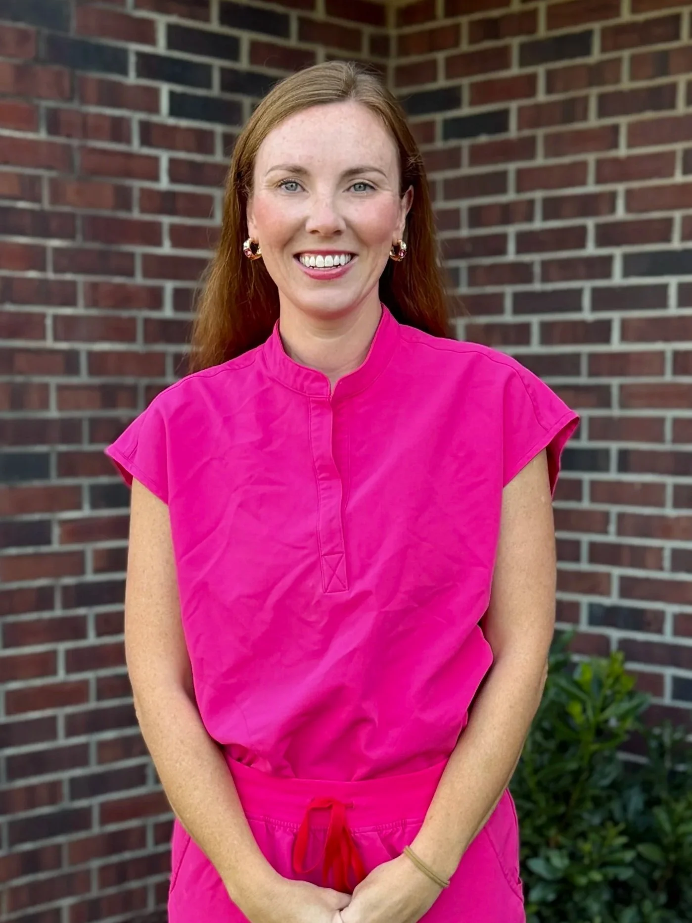 A smiling woman with red hair, wearing a bright pink top and matching pants, standing in front of a brick wall with green plants at the bottom right corner.