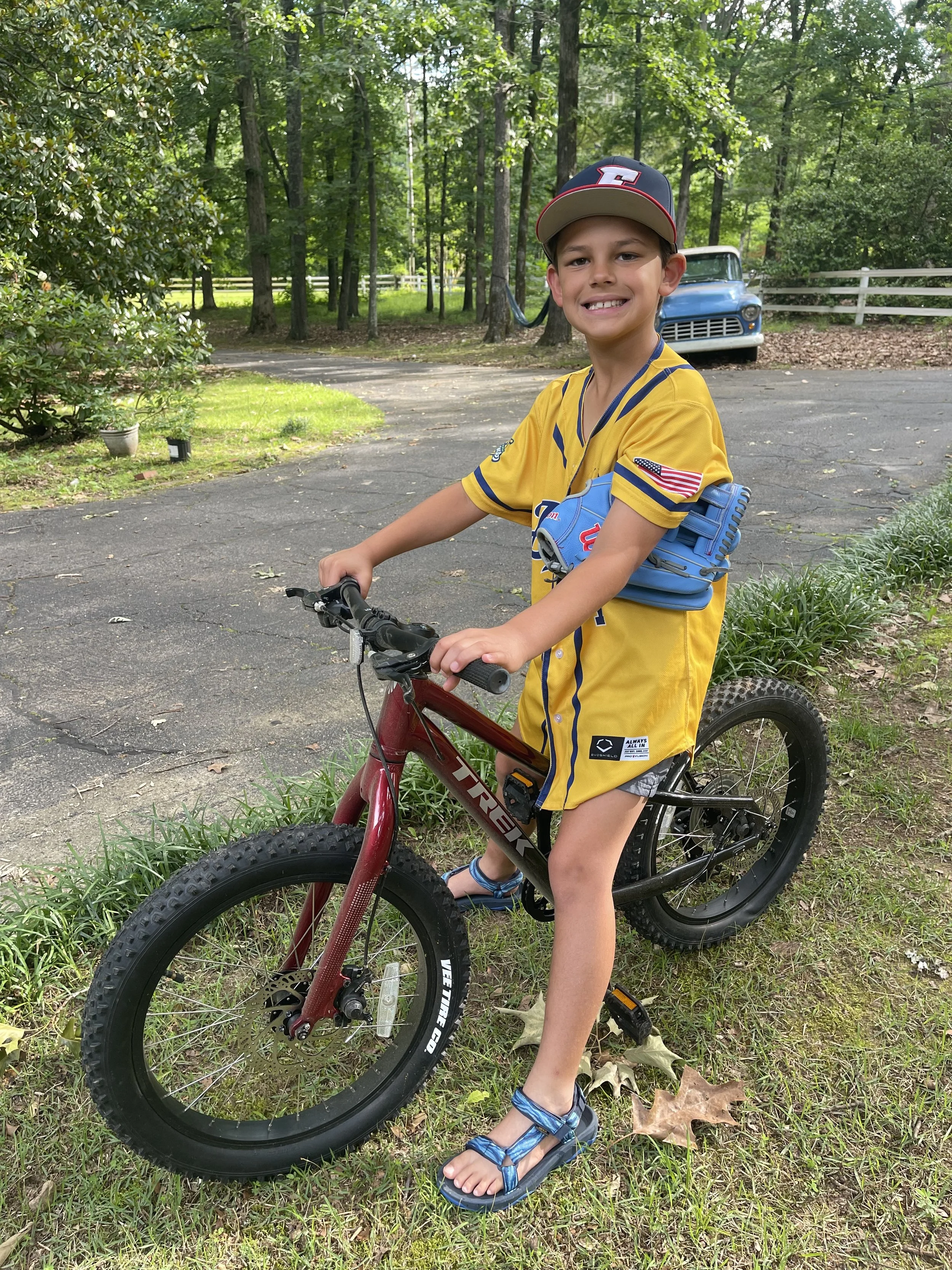 A young boy smiling while sitting on a mountain bike outdoors in a green, wooded backyard area with trees, a white fence, and a blue vintage truck in the background.