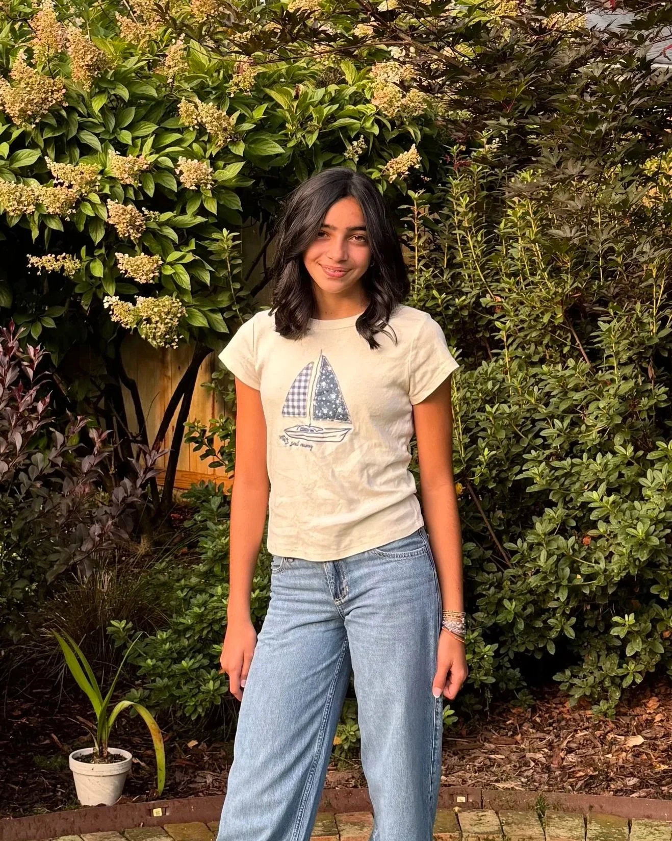 A young woman standing outdoors in front of lush green bushes and trees, smiling at the camera, wearing a beige t-shirt with a sailboat graphic and blue jeans.