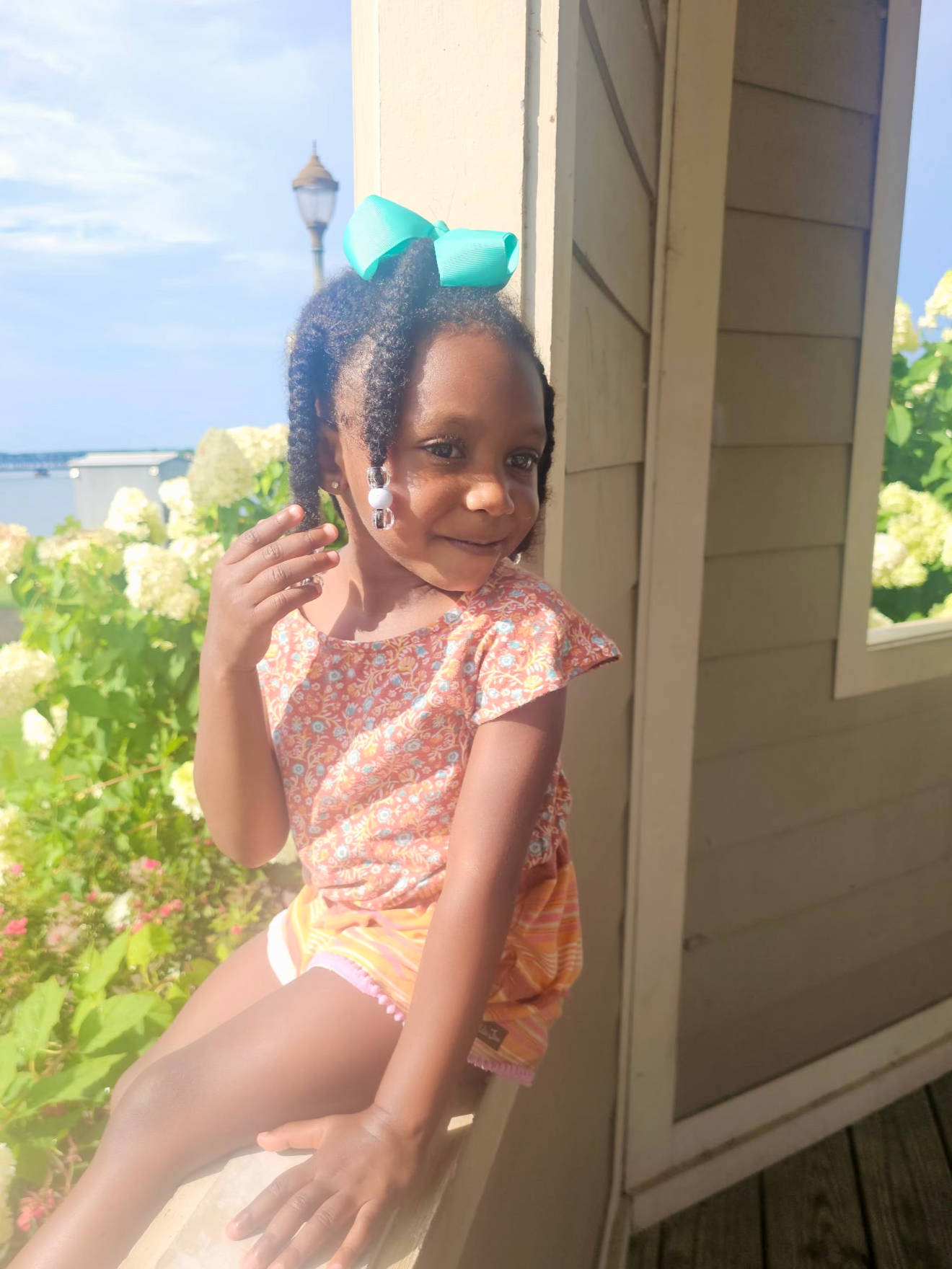 A young girl with dark curly hair wearing a large turquoise bow, earrings, and a floral orange top sitting outside on a wooden ledge near green bushes and hydrangeas, with a structure and a lamp post in the background.
