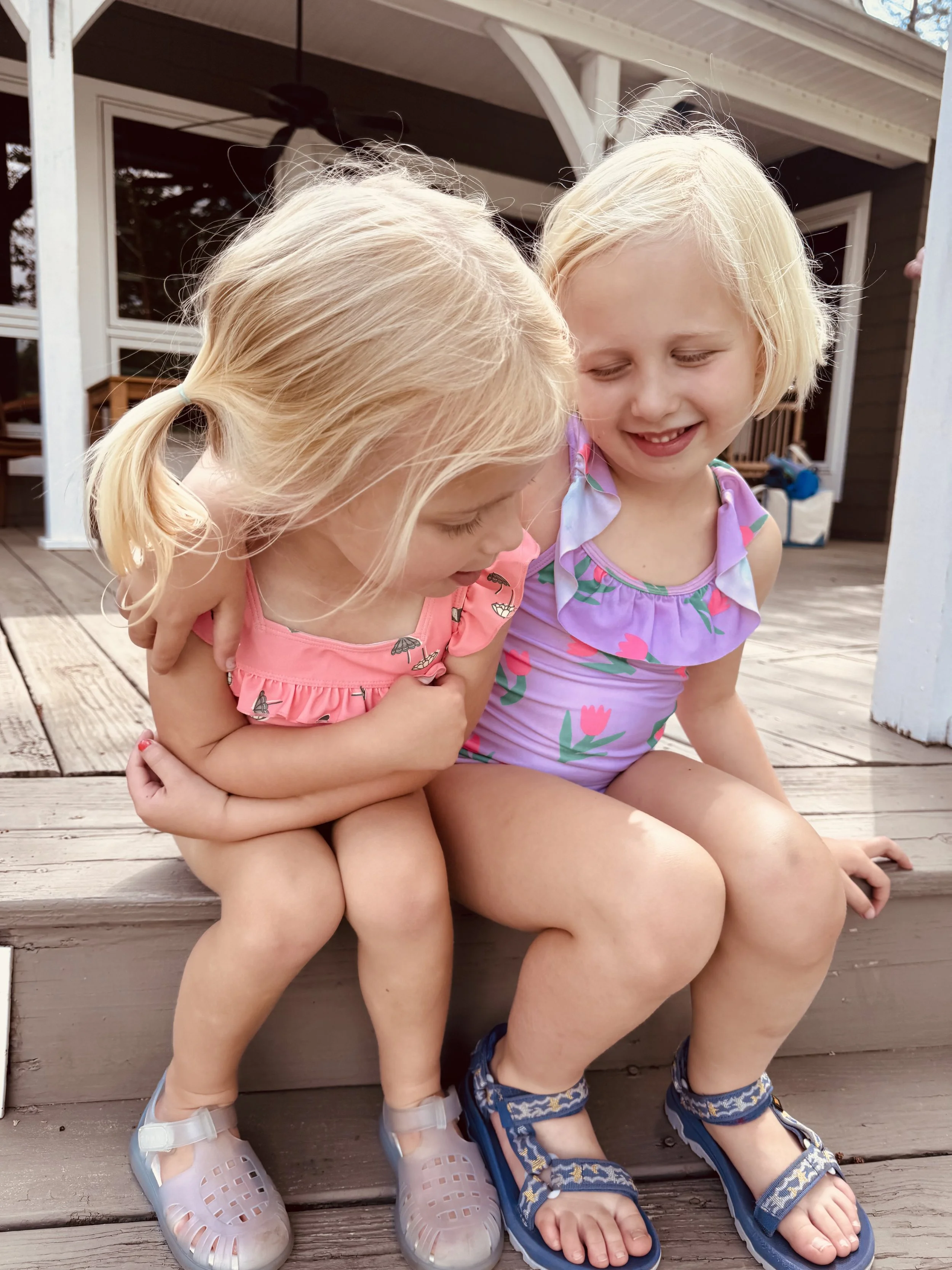 Two young blonde girls sitting closely on a wooden deck, smiling and enjoying each other's company, with one girl wearing a pink dress with butterfly patterns and the other a floral dress, both wearing sandals.