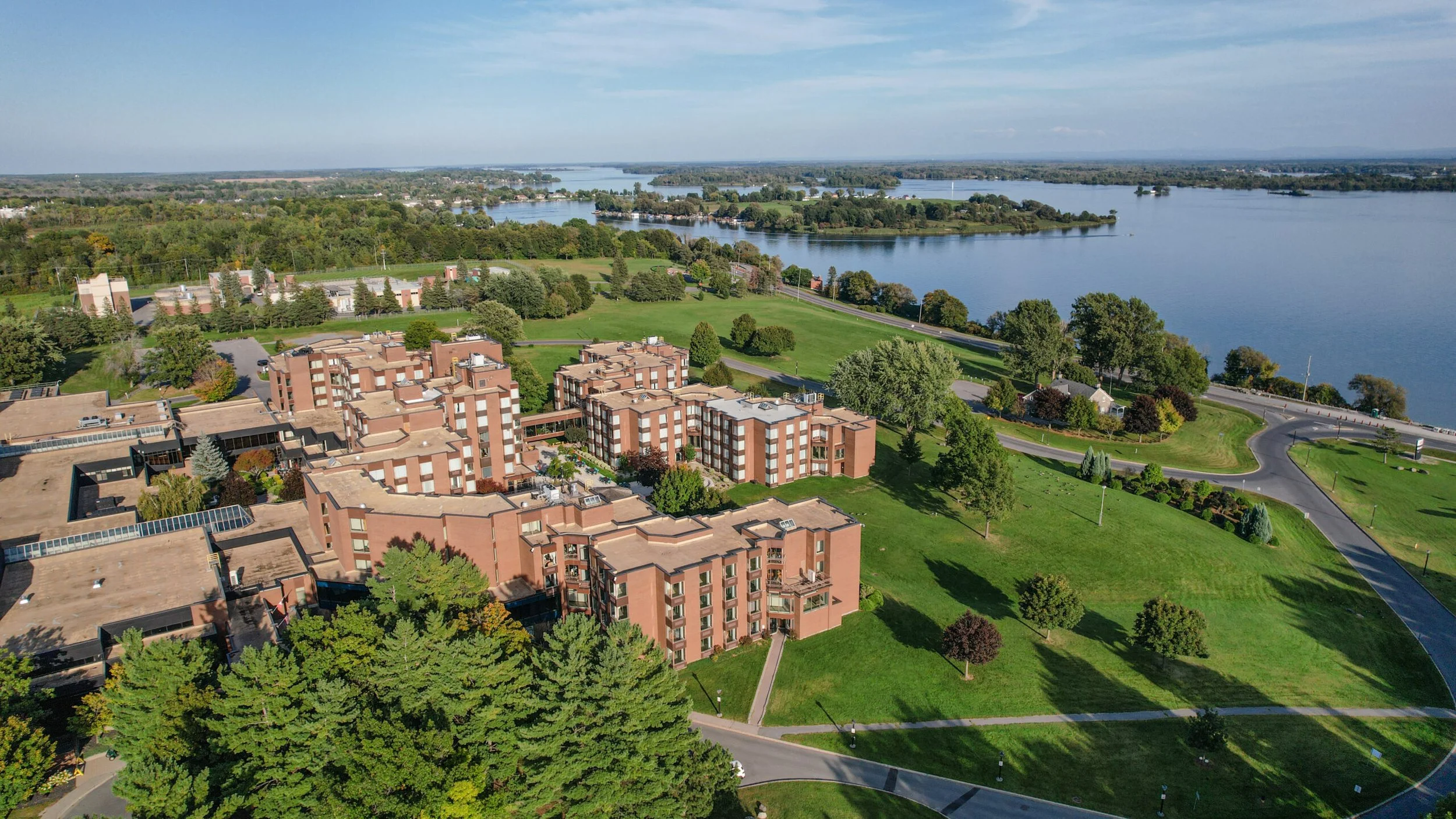An aerial view of a lakeside residential area with multiple apartment buildings surrounded by green trees, expansive lawns, and a large body of water with small islands in the background.