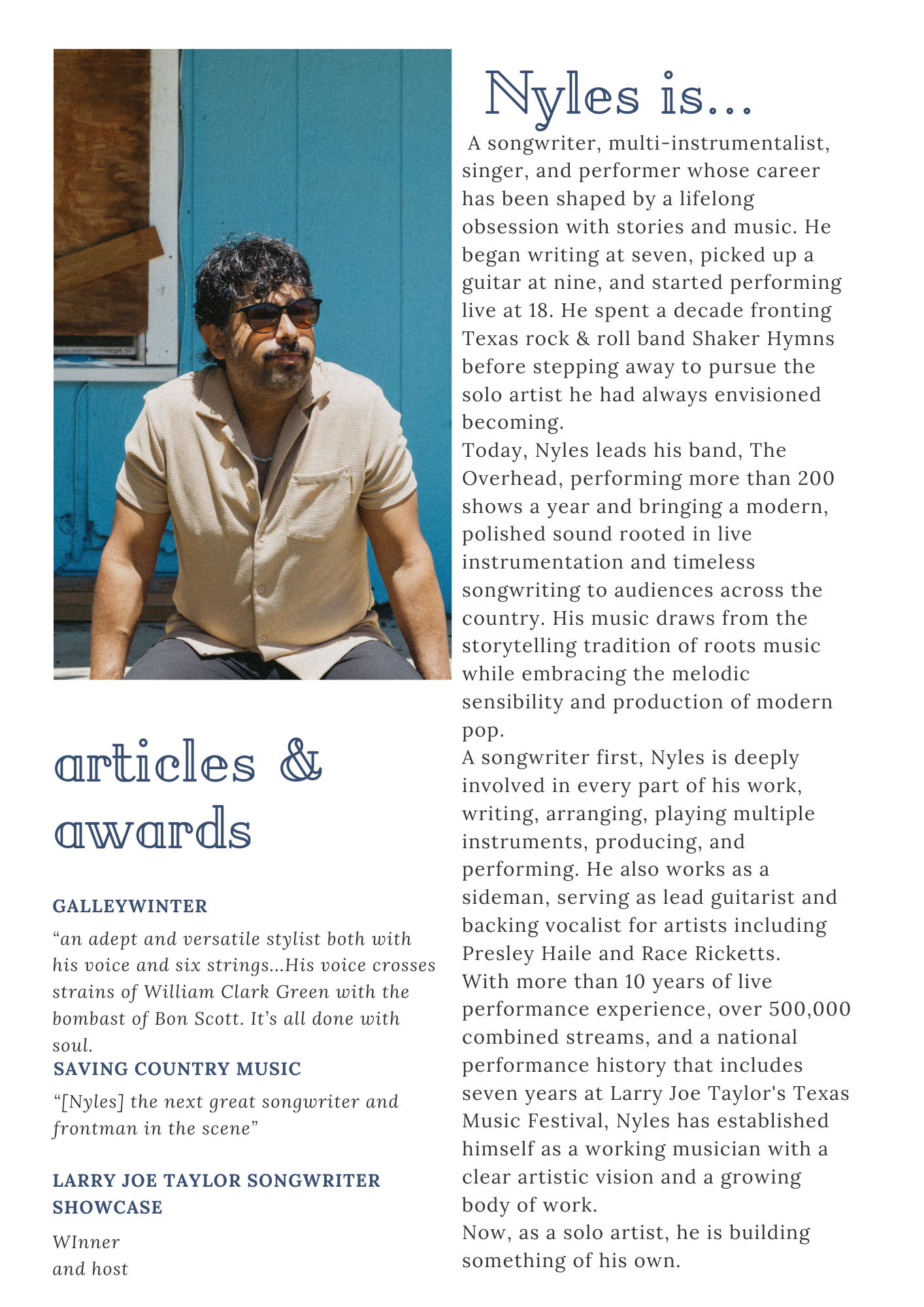 Photograph of Nyles, a man with dark curly hair, wearing sunglasses and a beige shirt with rolled-up sleeves, sitting outdoors in front of a blue wooden wall with a gravel or dirt surface.