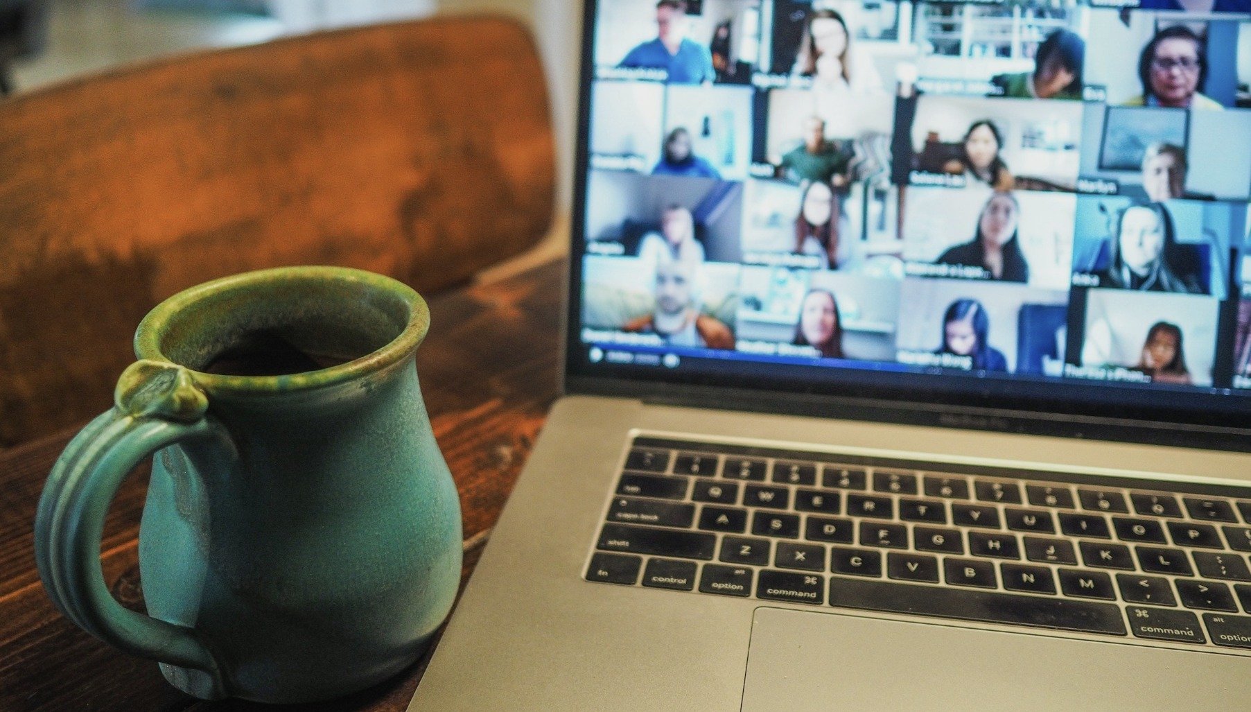A green ceramic cup filled with dark coffee on a wooden table next to a laptop showing a video conference with multiple participants.
