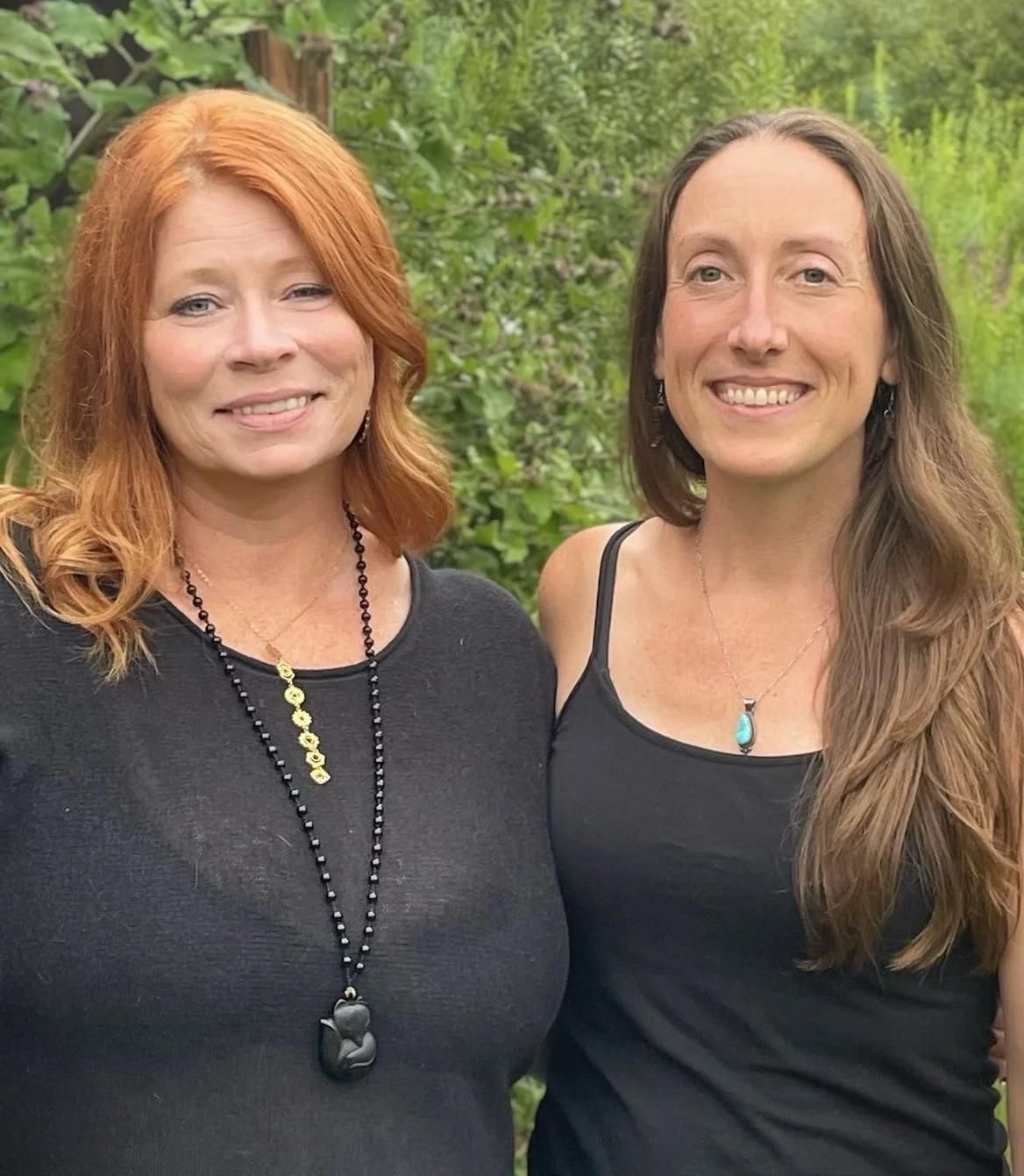 Two women standing outdoors surrounded by greenery, smiling at the camera, both wearing black tops and necklaces.