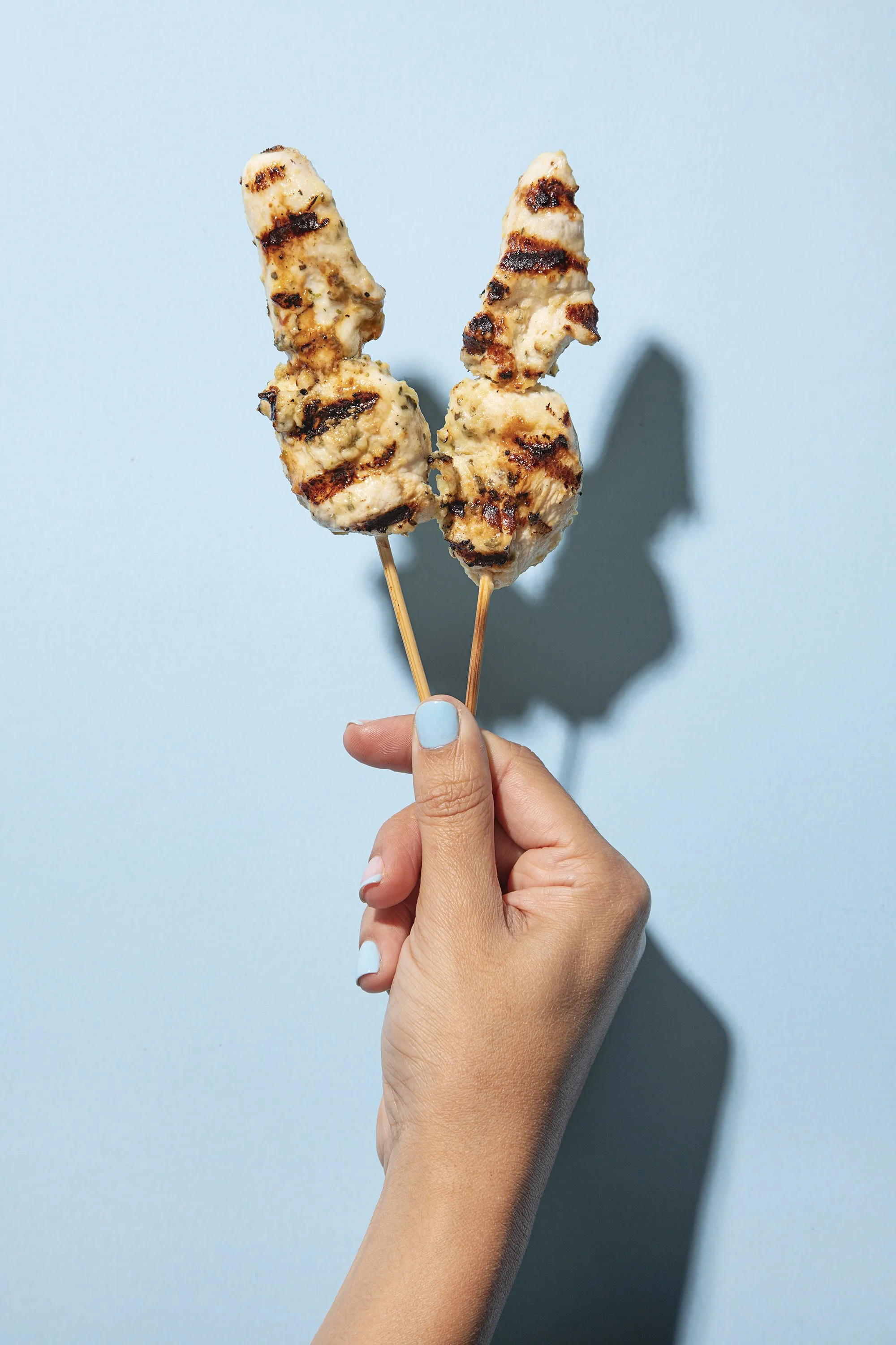 Two skewers of grilled chicken held by a hand with a light blue background.