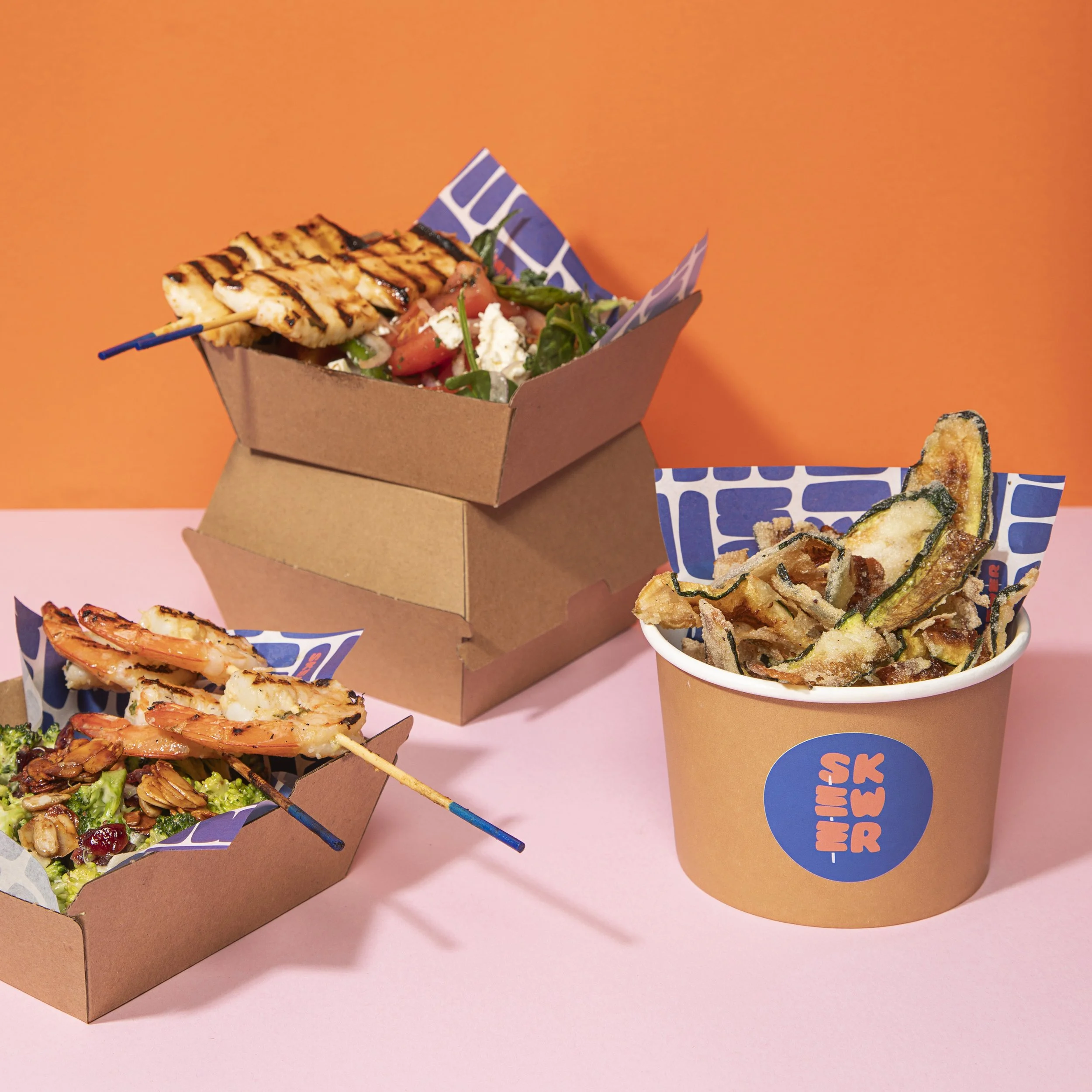 Takeout food containers with grilled skewers, zucchini chips, and salad against a coral orange background.