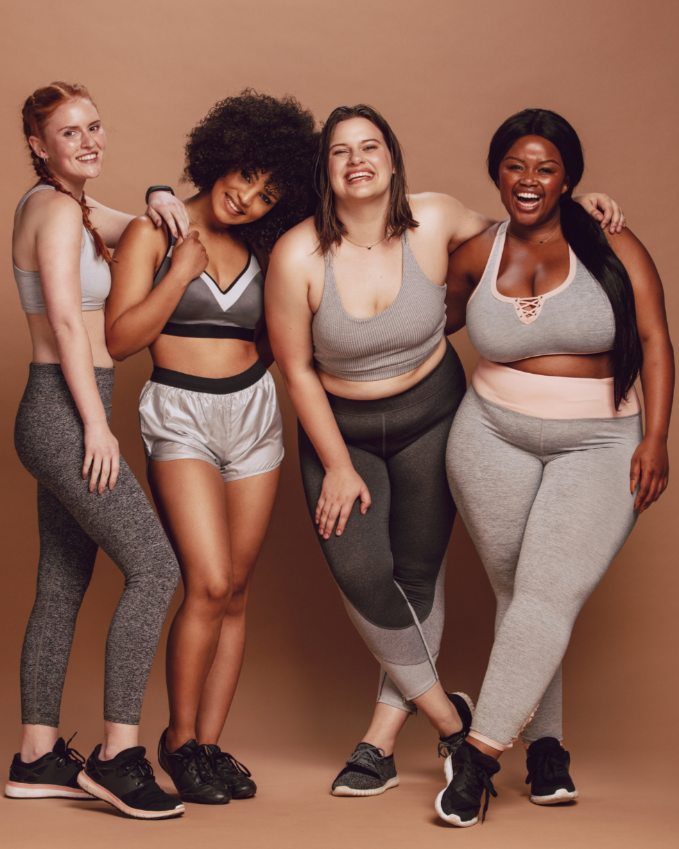 Four women in workout clothes smiling and posing together against a beige background.