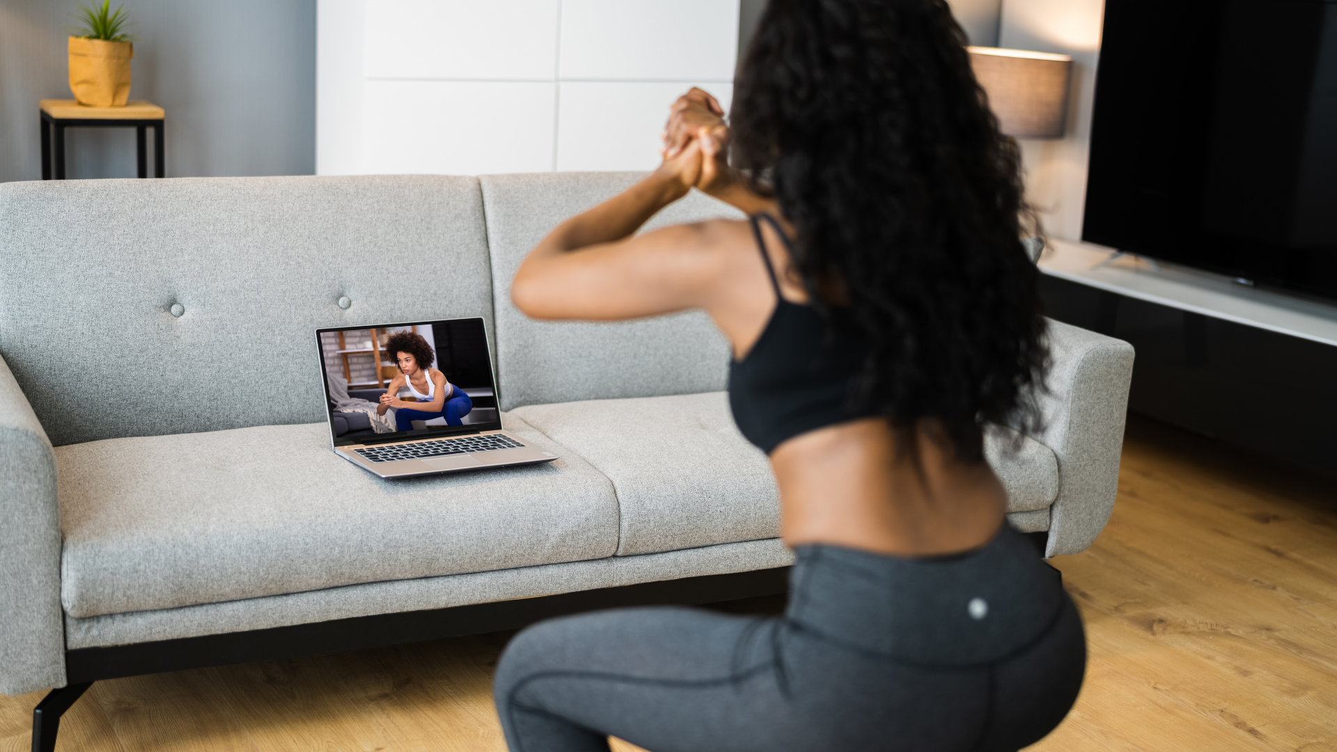 A woman with curly dark hair, wearing a black sports bra and leggings, is doing a squat exercise in front of a laptop on a couch. The laptop screen shows another woman in a workout pose, also in a black sports bra and blue leggings, participating in a virtual workout session in a living room.