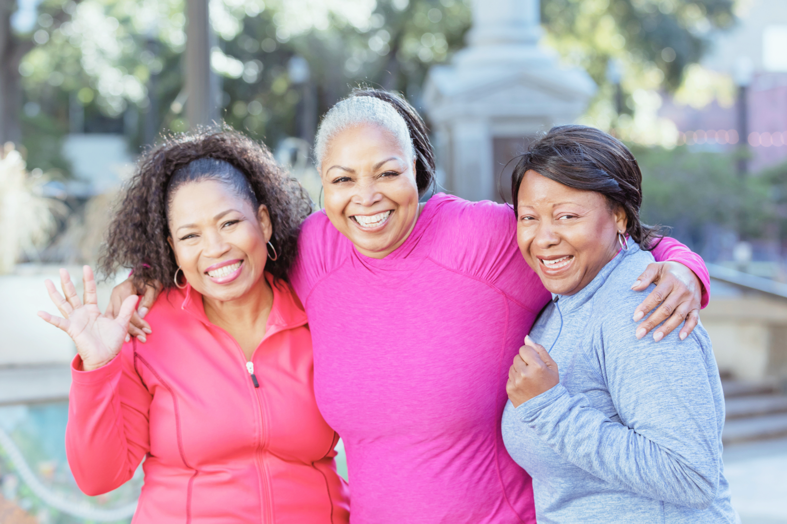 Three women smiling and hugging outdoors in a park, two of them waving, wearing bright activewear.