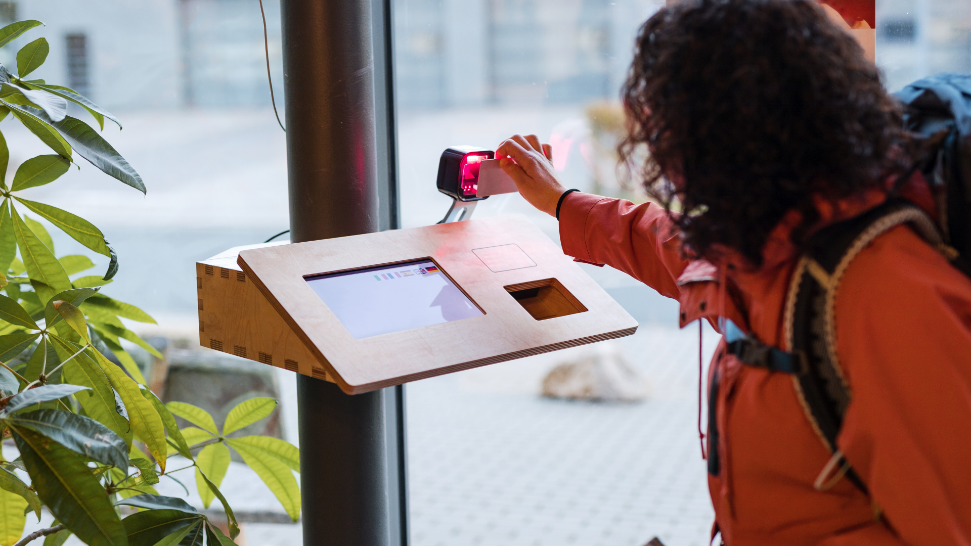 A person using a contactless payment terminal to make a transaction at a table with a display screen, seated near a window with greenery outside.