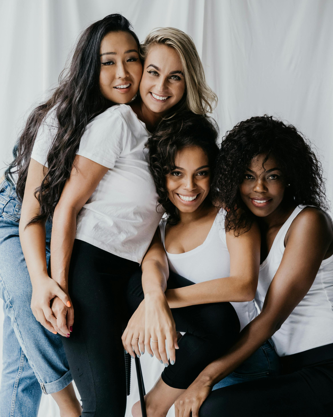 Four women with diverse ethnicities smiling and sitting close together, dressed in casual white tops and black pants or jeans, against a white background.