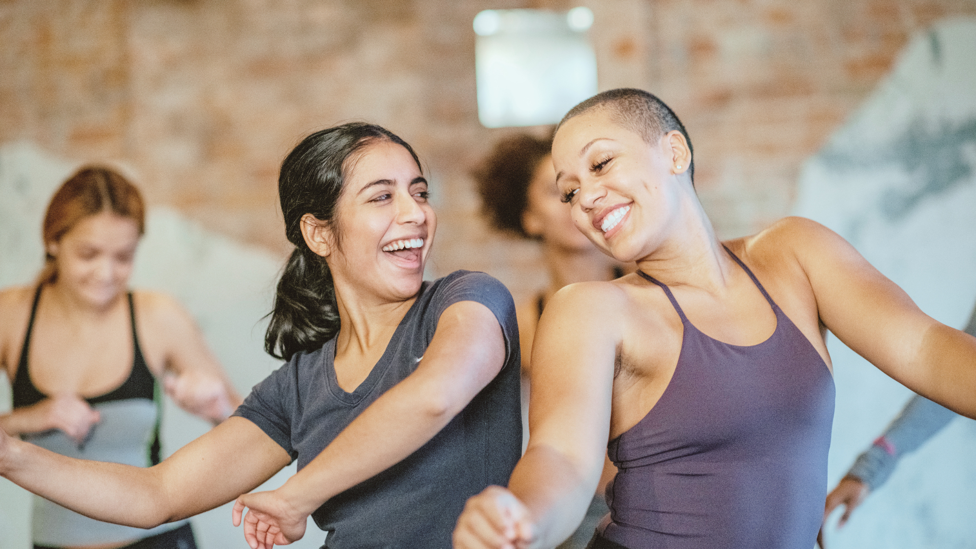 Two women smiling and stretching during an exercise class in a gym, with other women in the background.