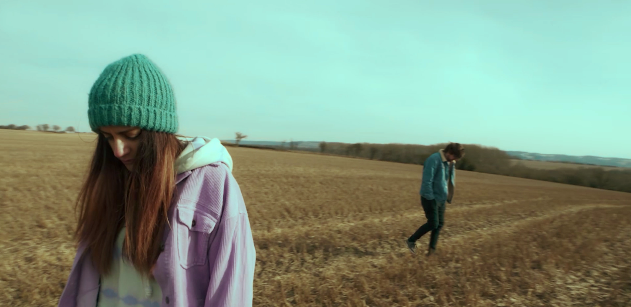 Two people walking in a rural field during daytime, one with a teal beanie and pink jacket, and the other in a blue jacket and black pants.