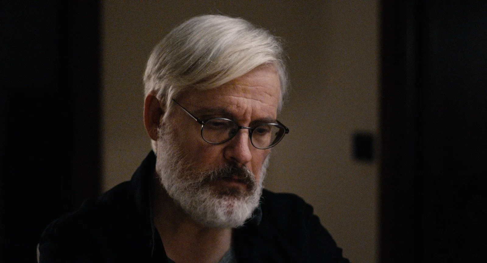 Close-up of an older man with gray hair, beard, and glasses, looking down with a serious expression.