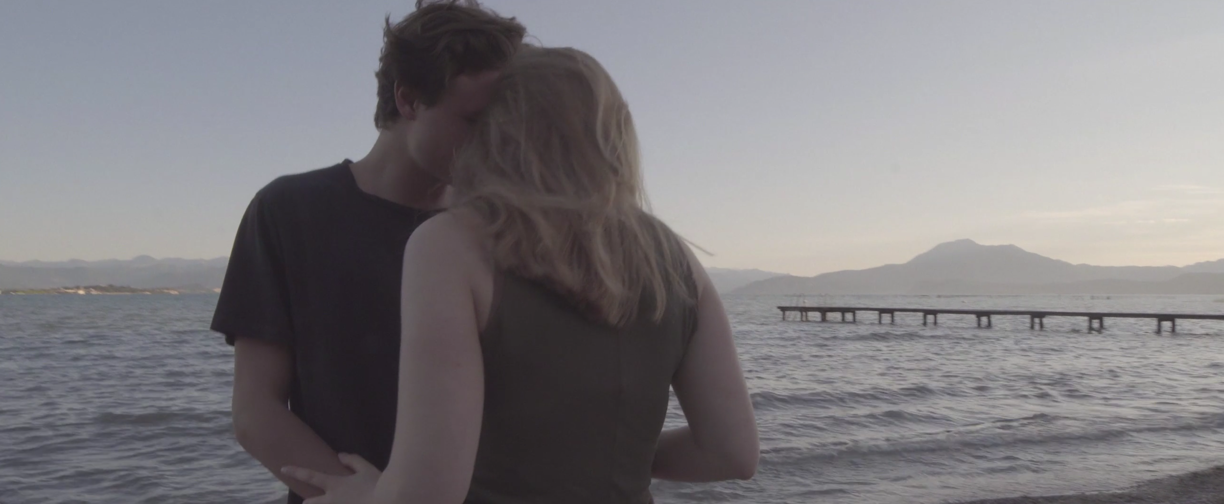 A young couple kissing by the water with mountains and a pier in the background at sunset.