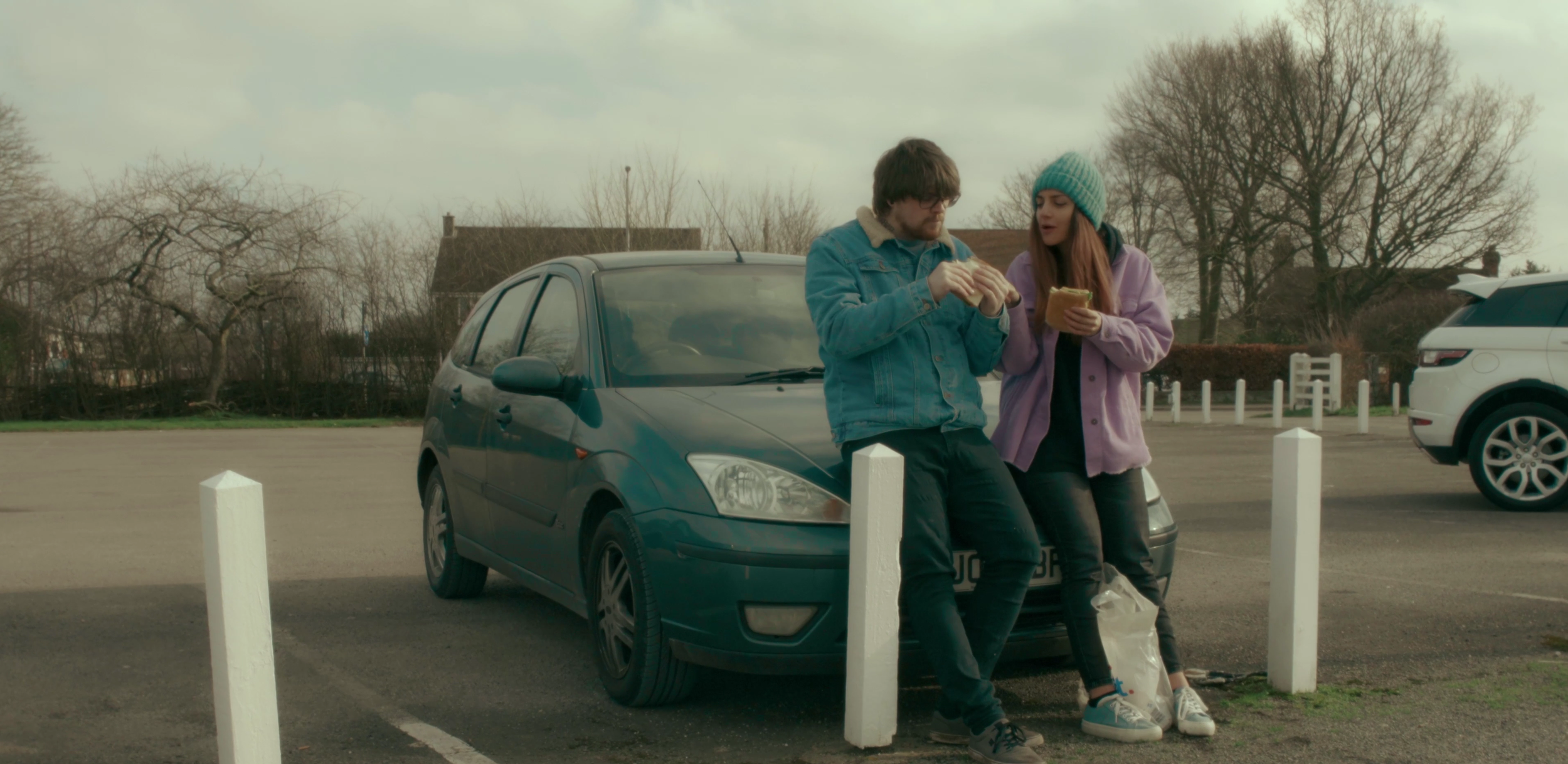A young man and woman sitting on the hood of a blue car in a parking lot, eating sandwiches and talking. The woman is wearing a purple jacket and a teal beanie, while the man is wearing glasses and a blue denim jacket. There are trees and other parked cars in the background.