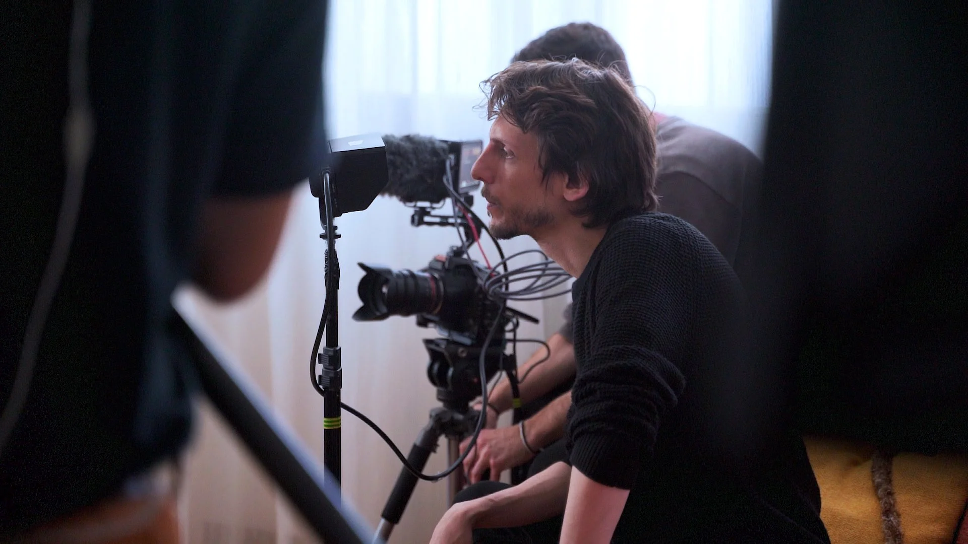 Man with wavy brown hair and a black shirt operating a professional film camera indoors, with another person partially visible behind him and a woman in the background, during daytime.