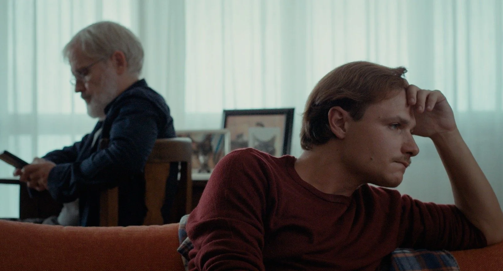 A young man sits on an orange couch with his hand on his forehead, appearing frustrated or deep in thought, while an older man with glasses and a gray beard stands in the background looking at his phone in a softly lit room.