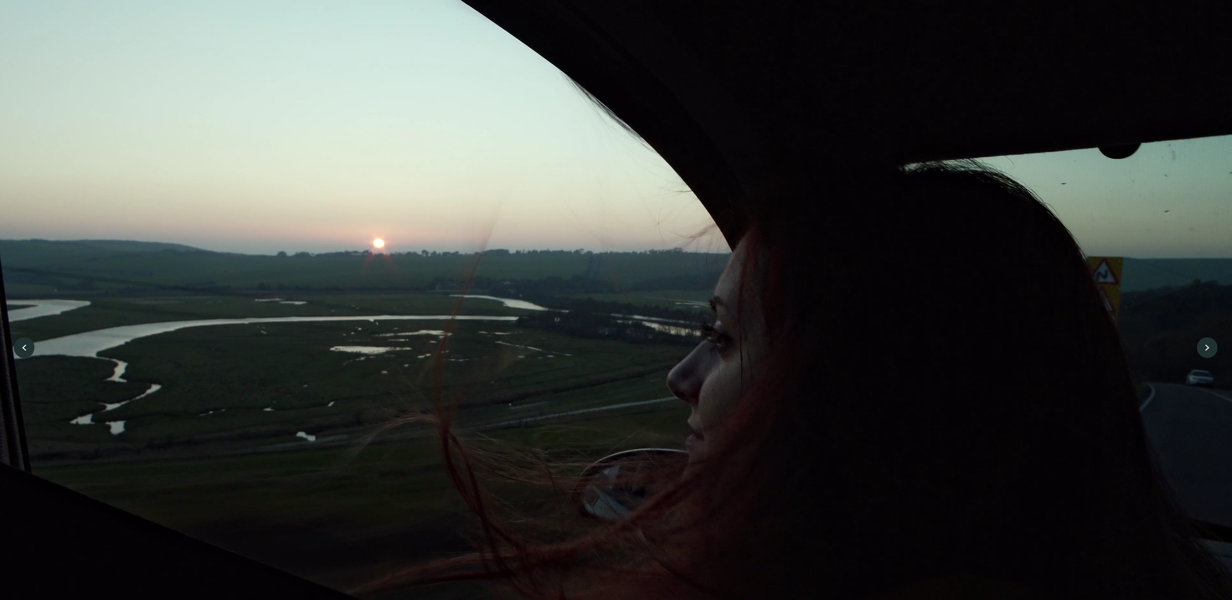 Person with long hair sitting in a vehicle, looking out the window at a sunset over a winding river and rolling hills.
