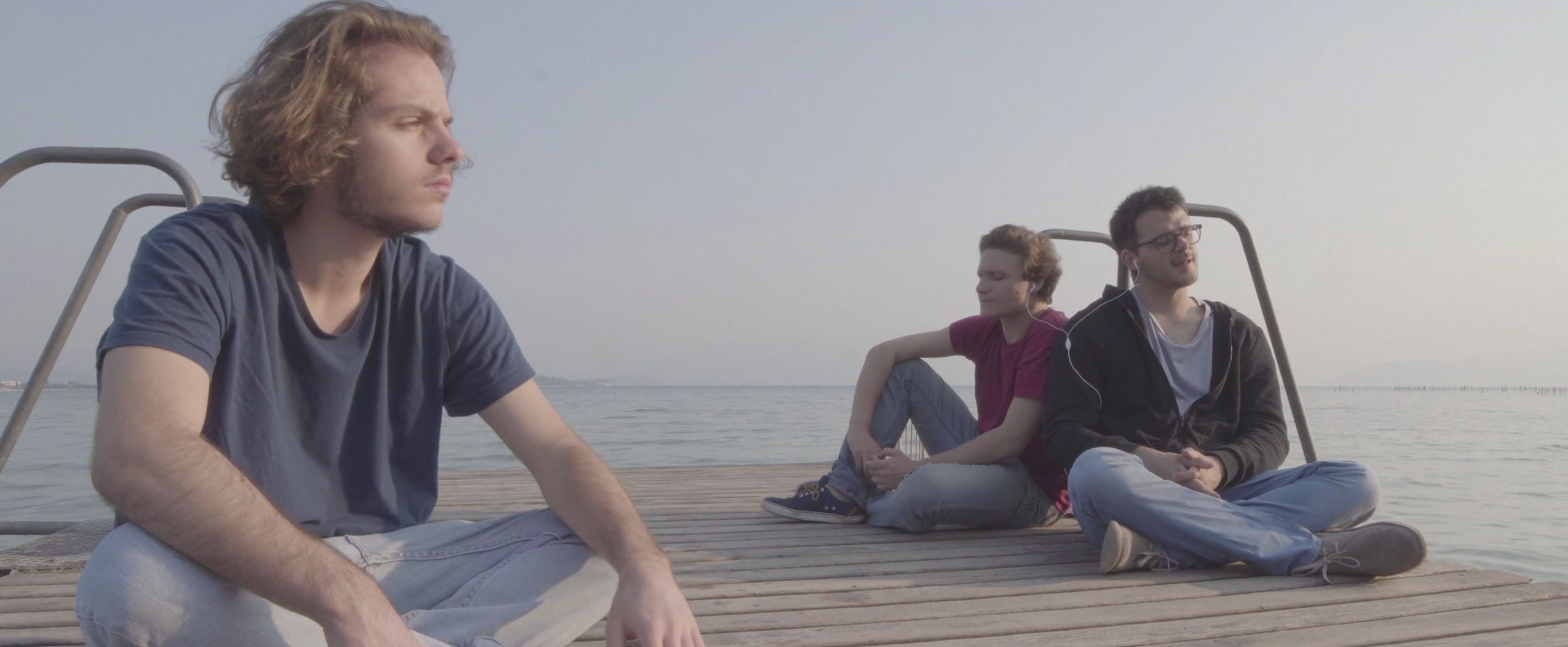 Three young men sitting on a wooden dock by the water, facing different directions. The first man is in the foreground with curly hair, wearing a blue t-shirt and light pants. The second man, in the background, has short hair, wearing a red t-shirt, and is listening to music with headphones. The third man has glasses, dark hair, and a jacket, and is also listening to music with earphones.