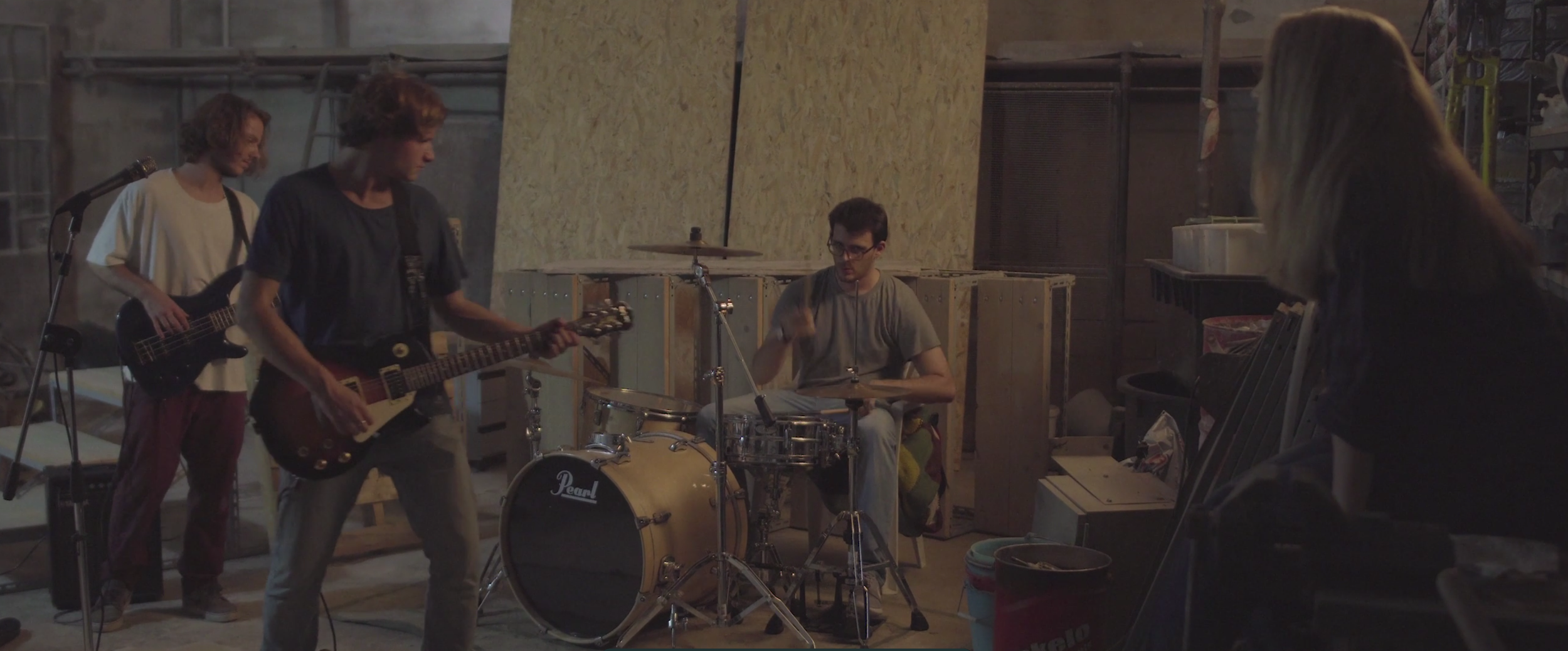Four young people rehearsing a band in a garage with musical instruments, including guitars, drums, and a bass guitar.