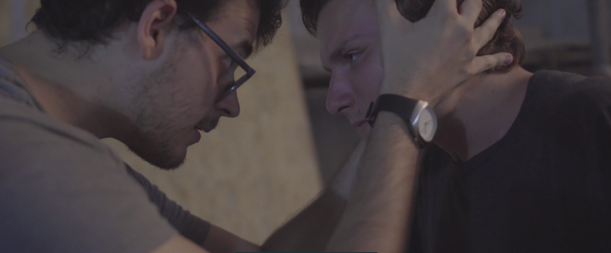 Two young men face each other closely, one with glasses and a beard, and the other with short hair and a watch, appearing to share an intense emotional moment.