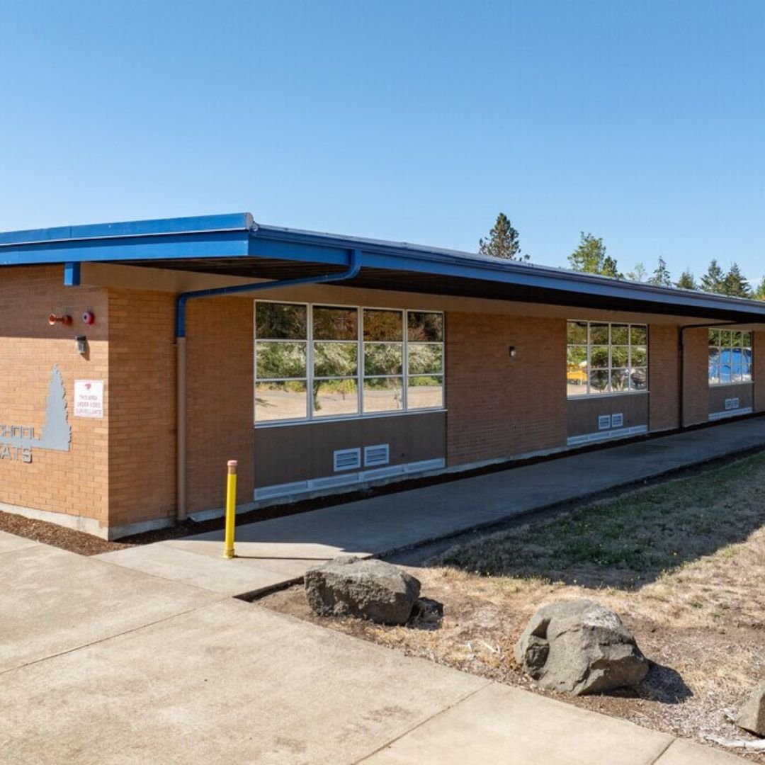 A single-story brick building with large windows and a blue roof, outdoors on a sunny day, with rocks and sidewalk in the foreground.