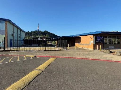 Glide School District Parking lot area with a yellow curb, a small brick building, and a larger blue industrial building, under a clear blue sky.