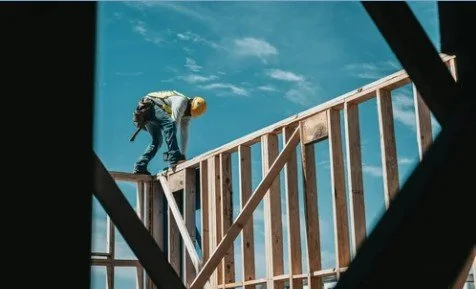 A construction worker wearing a yellow hard hat and safety vest is working on a wooden frame of a building under construction, viewed through a window or opening.