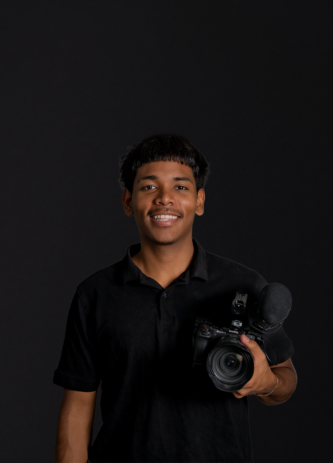 Young man smiling and holding a professional camera with a microphone attached, standing against a dark background.