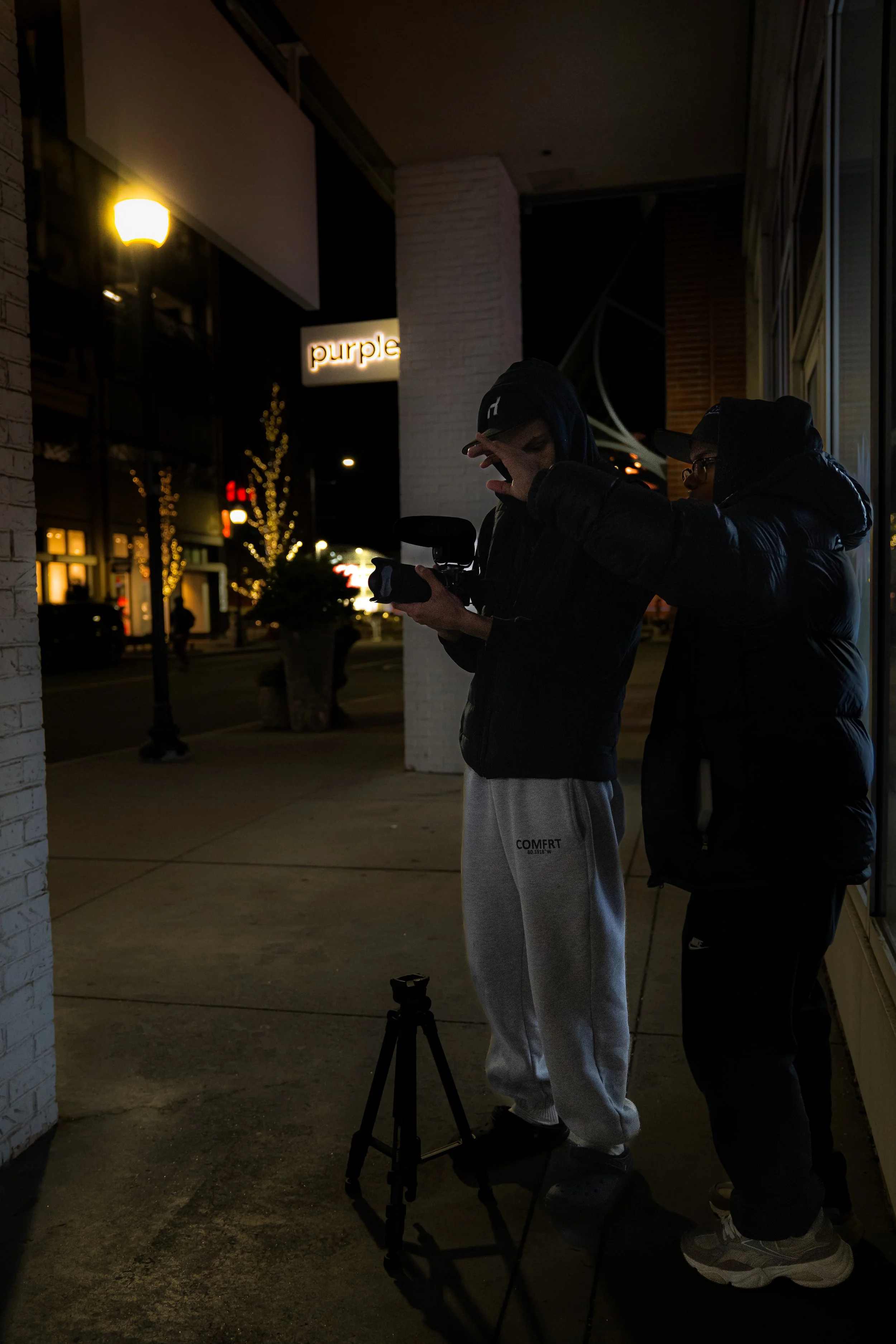 Two young men standing outside at night, one looking at a camera on a tripod, the other adjusting his hood, with a lit sign reading 'purple' in the background and string lights on trees.