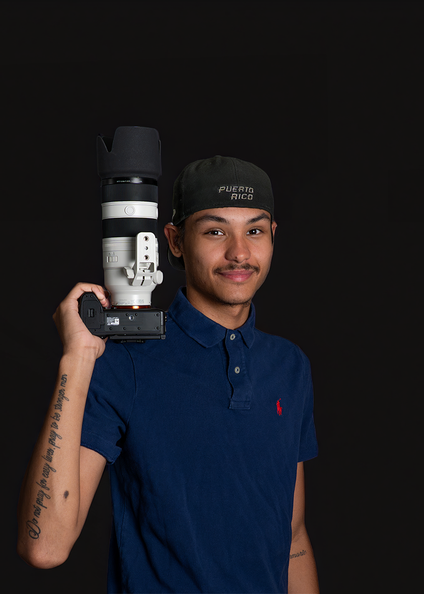 Young man with a mustache wearing a black Puerto Rico baseball cap and a navy blue polo shirt, holding a large camera with a telephoto lens on his shoulder, standing against a black background.