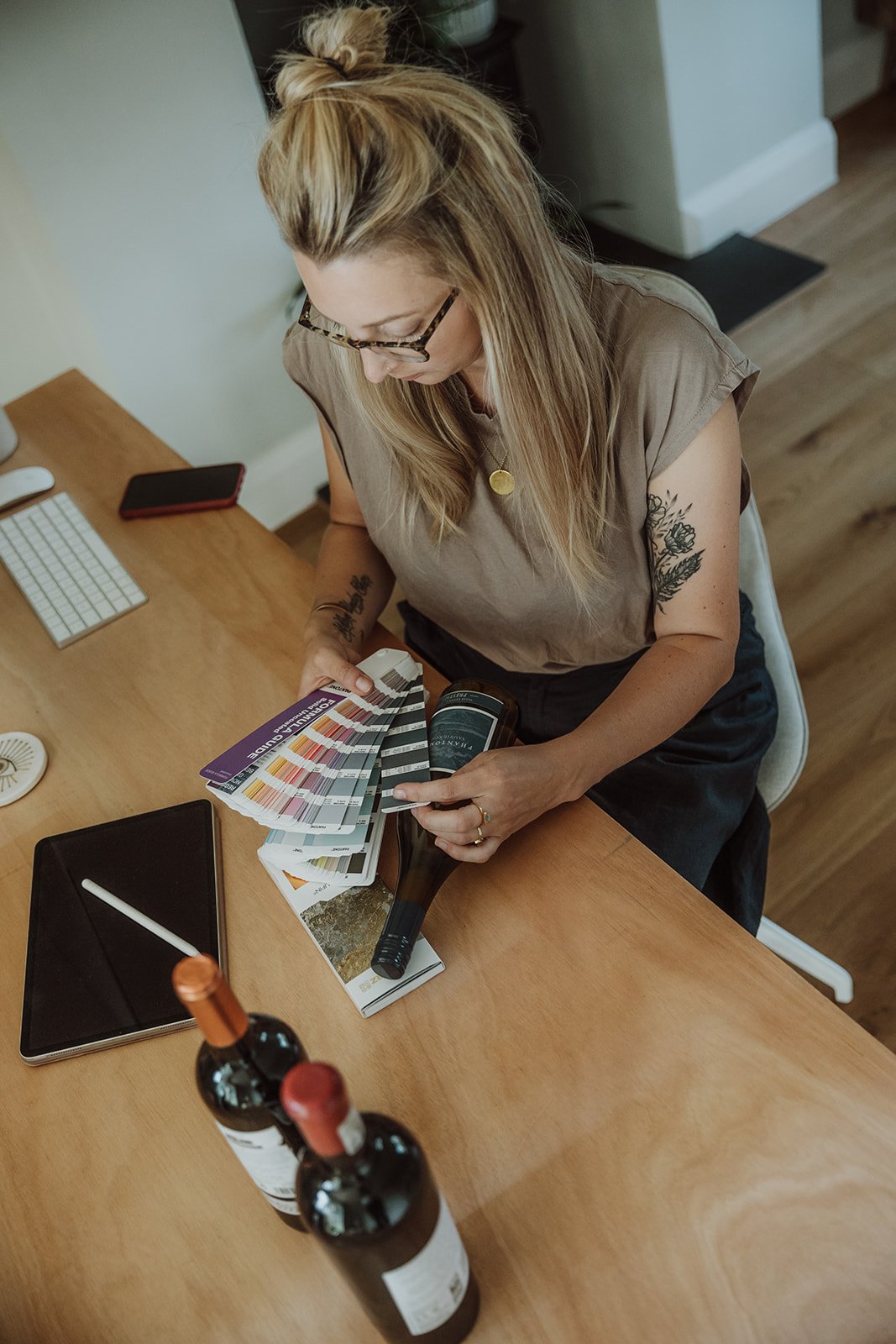 Woman sitting at a desk examining paint color swatches, with a microphone and bottles of wine on the table.