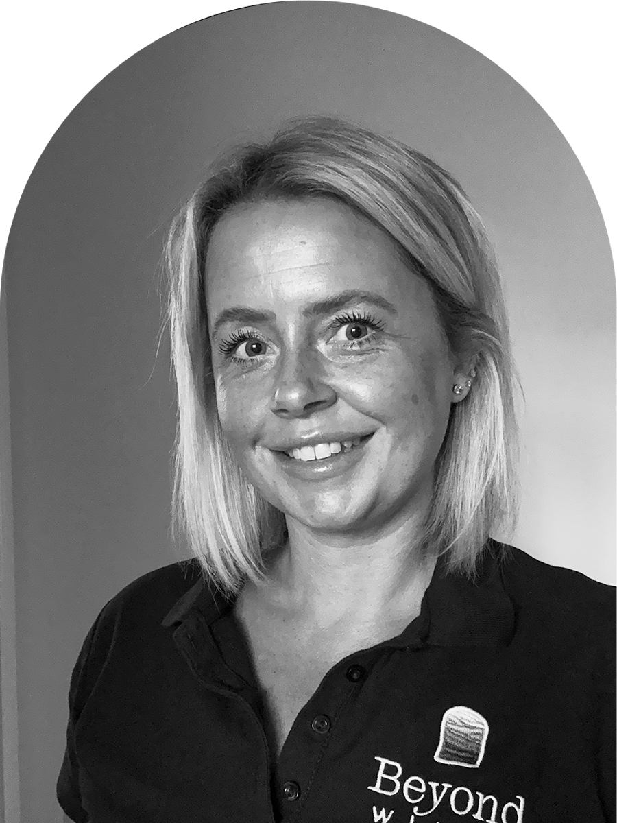 A black-and-white photo of a woman with shoulder-length blonde hair, smiling, wearing earrings and a collared polo shirt with the logo and text 'Beyond' on it.
