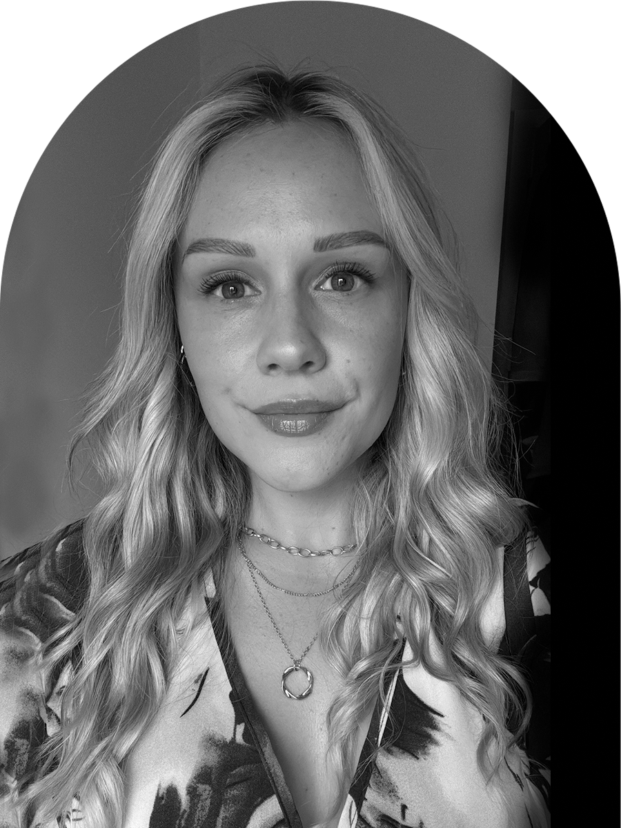 A black-and-white portrait of a woman with wavy blonde hair, wearing layered necklaces and a patterned top, smiling softly.