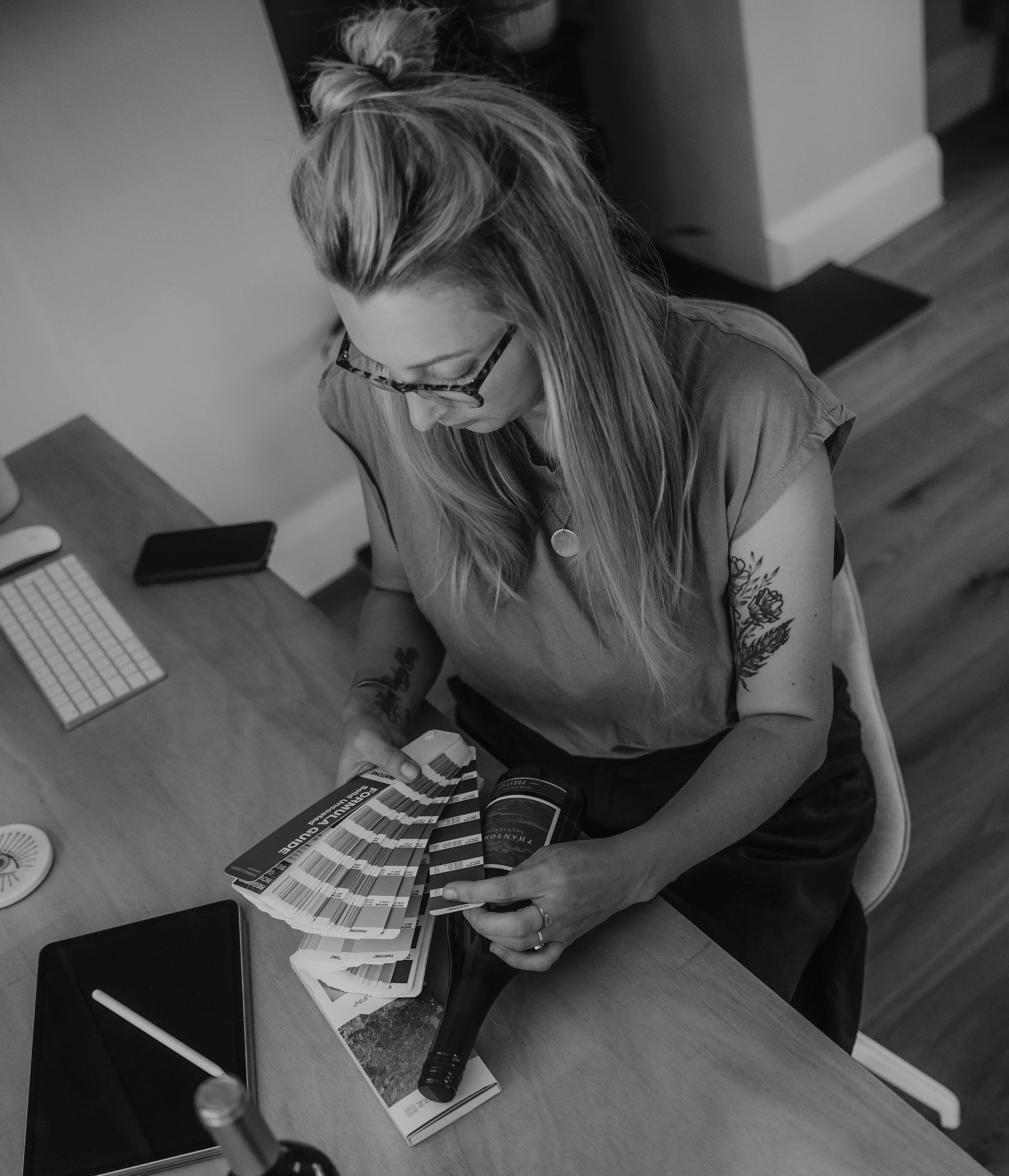 A woman with glasses and tattoos on her arm is holding a stack of color swatches or sample cards at a table. Various electronic devices, including a tablet, a smartphone, and a digital stylus, are on the table.