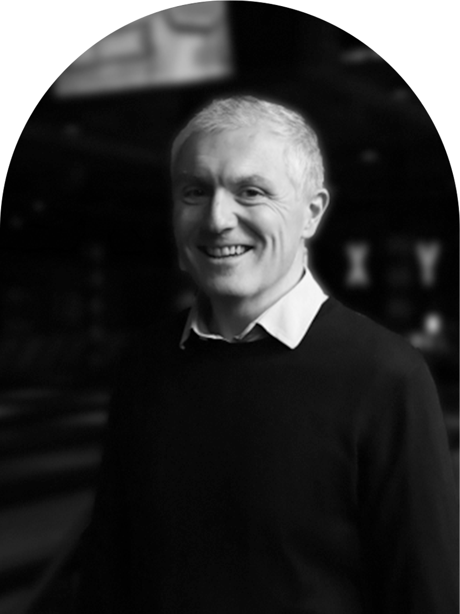 Black and white photo of a smiling man with short, light-colored hair, wearing a collared shirt and a dark sweater, standing indoors.