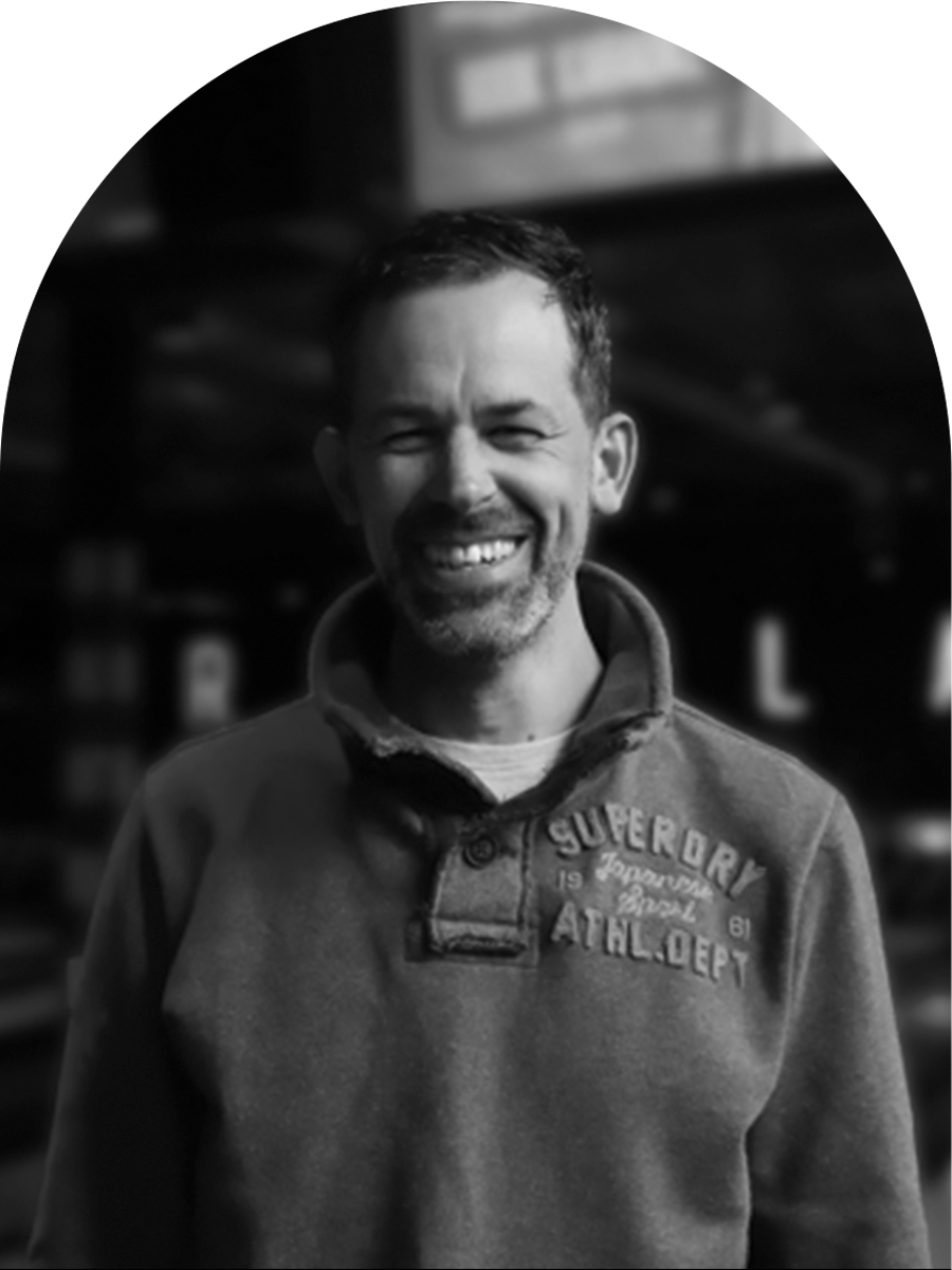 A smiling man with short dark hair and facial hair, wearing a sweatshirt with embroidered text, standing indoors.