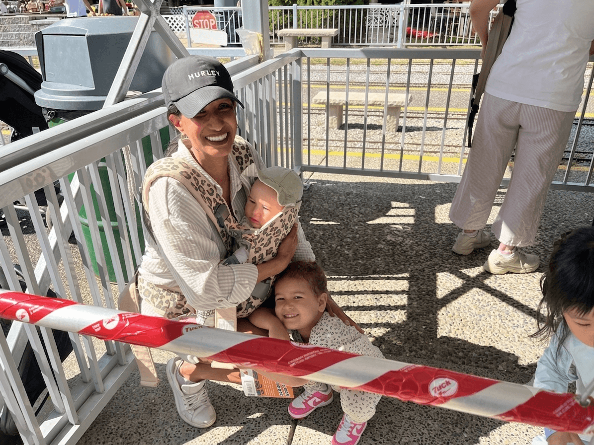 Mother crouching down and holding a baby strapped to her chest. A toddler is hugging mothers knees while waiting for train ride at Burnaby Central Railway