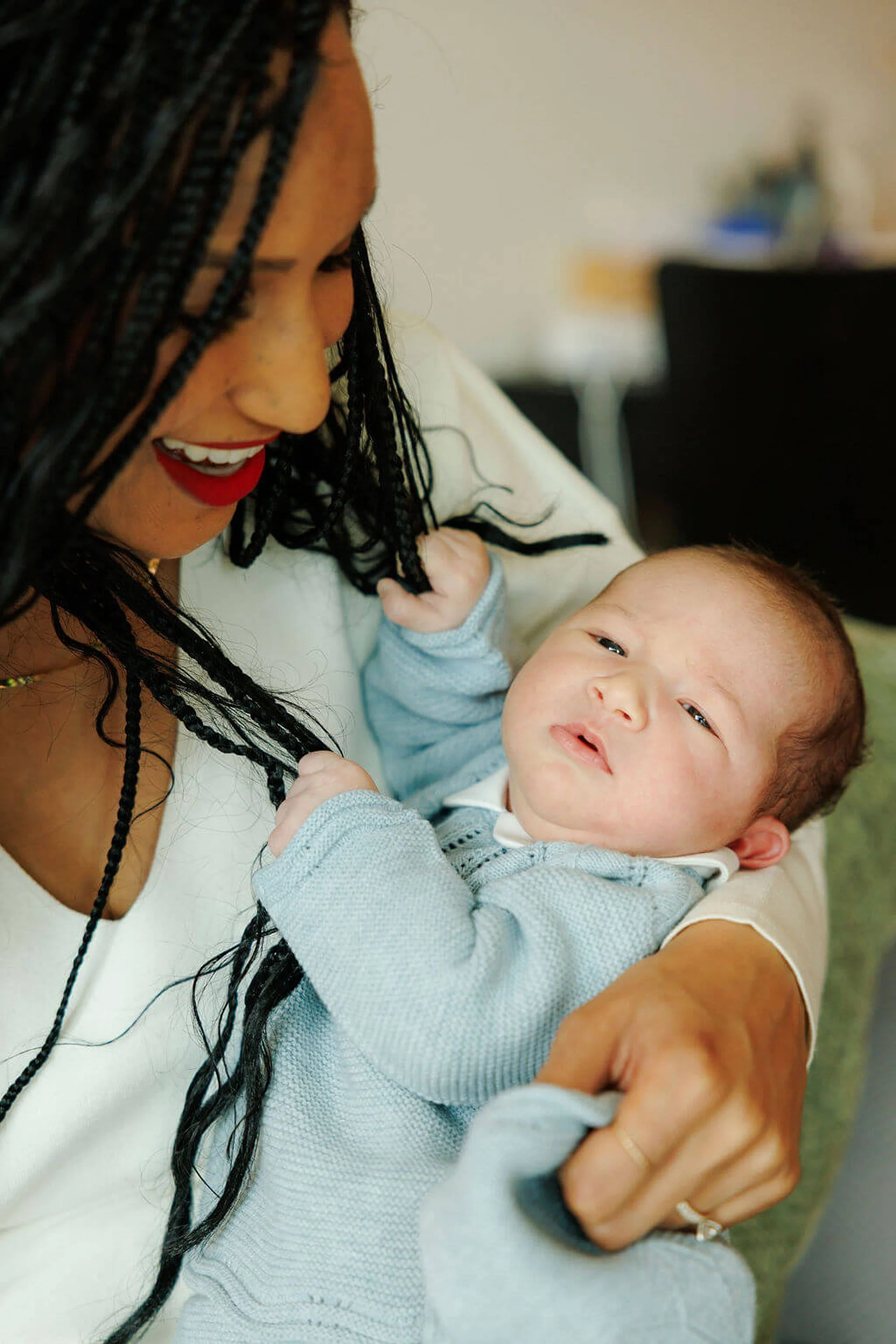 A woman with long braided hair and red lipstick holding and looking at a young child with a curious expression.